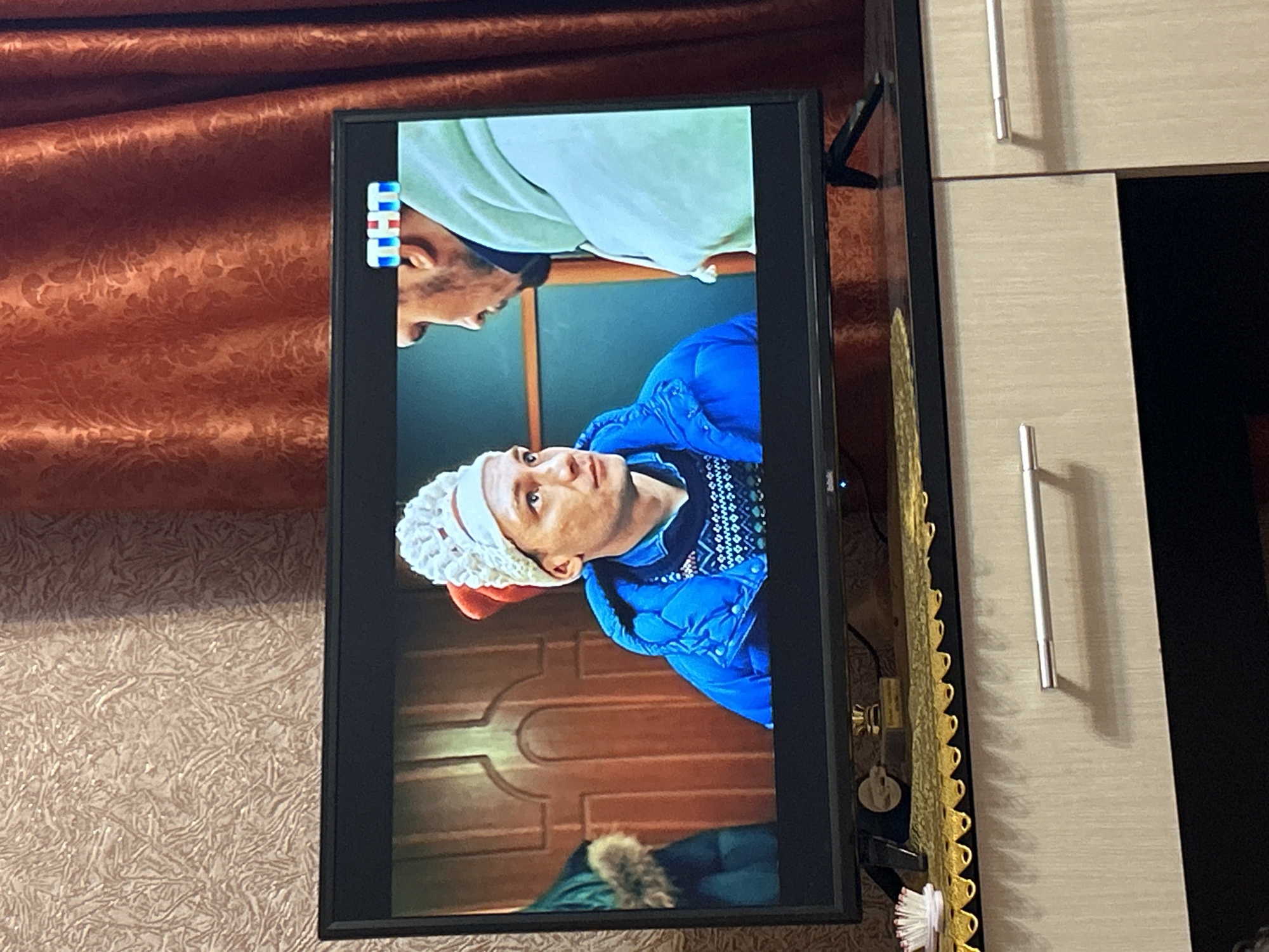 The image shows a flat-screen television displaying a scene from a television show. Two men are visible on the screen. The man on the left is wearing a blue puffer jacket and a white knitted hat with a red pom-pom. He has fair skin and light-colored hair. The man on the right is wearing a light green hooded sweatshirt and is facing the other man. The television is placed on top of a wooden cabinet with two drawers, each with a silver handle. A patterned curtain is visible in the background, to the right of the television. The wall behind the television has textured wallpaper. A small trophy is visible on the cabinet shelf behind the television. The scene appears to be indoors. The image does not contain information indicating the location in Gelendzhik, Russia.