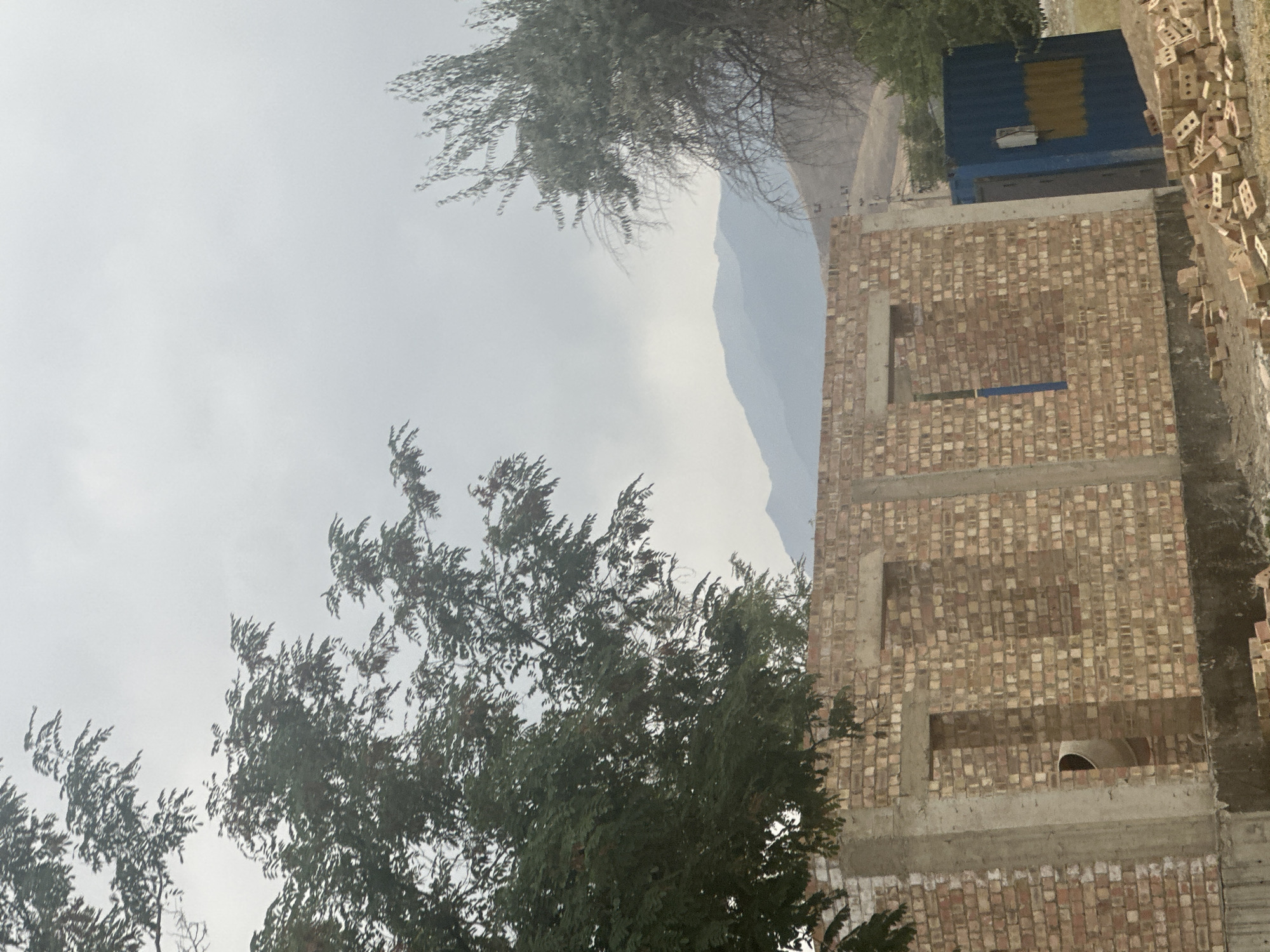 An overcast day in Алмалуу, Kyrgyzstan, shows a construction site. In the foreground, a brick building under construction has concrete pillars and openings for windows and a door. Piles of loose bricks are scattered on the ground in front of it. To the right, a blue shipping container with a yellow stripe is partially visible. In the background, several trees, one with dark leaves and red berries, and another with bare branches, frame the scene. Behind the trees, layered mountains recede into the hazy sky.