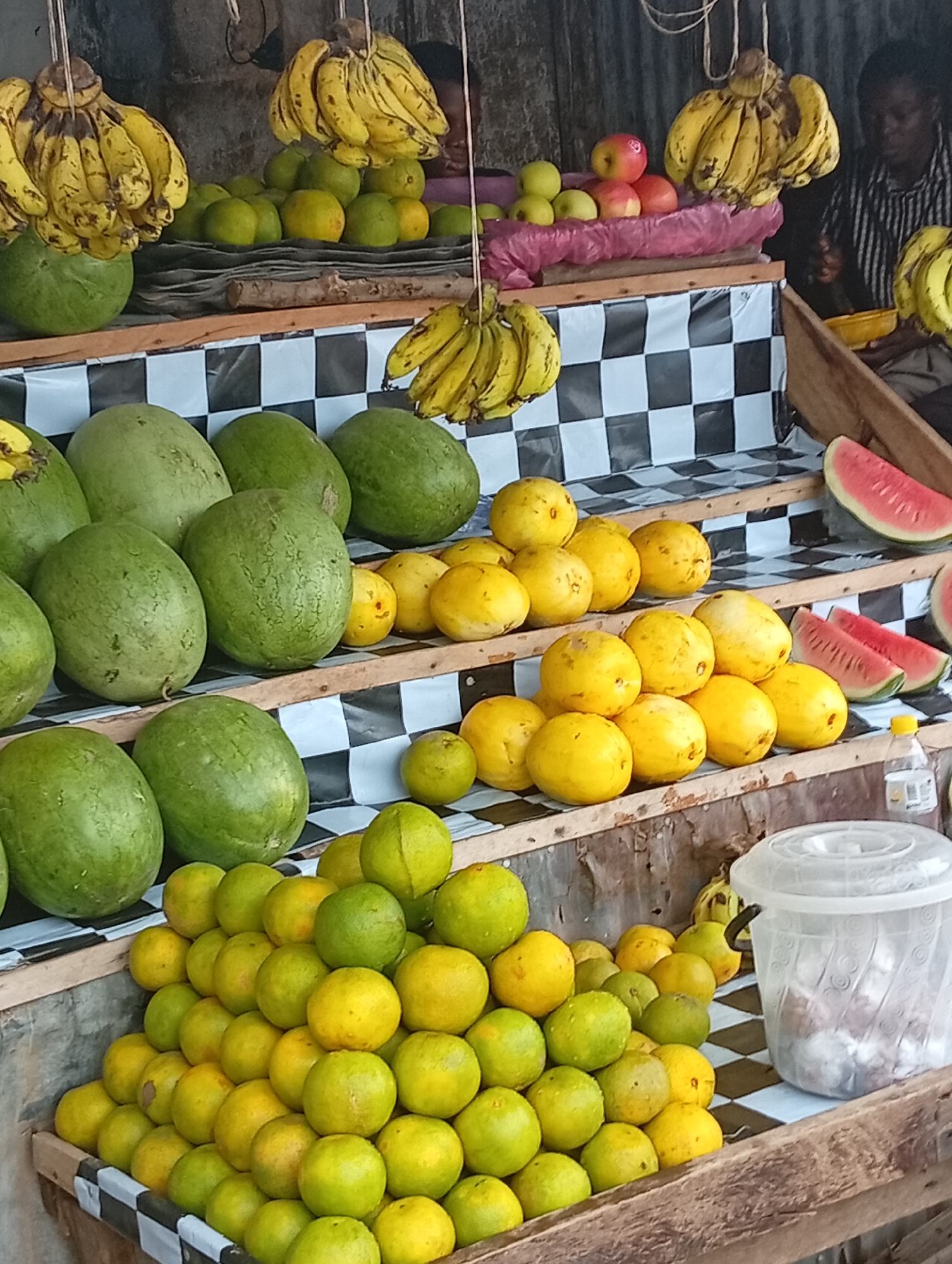 A street market stall in Jos, Nigeria, displays an abundant variety of fresh fruits for sale. The stall features multiple tiered shelves constructed from wood and covered with a black and white checkered fabric.
Prominently featured are large, green watermelons, arranged in rows on the higher shelves. Below them, a significant quantity of round, yellow-orange fruits, likely oranges or tangerines, are piled high in a wooden crate. Smaller piles of these yellow-orange fruits are also visible on other shelves, interspersed with some green citrus fruits.
Further up, bunches of yellow bananas are displayed, some hanging from above and others resting on shelves. Red apples are also visible on the top shelf, placed next to a bunch of bananas. Slices of watermelon are arranged on a wooden tray on the right side of the stall.
In the background, a person wearing a striped shirt is partially visible, seated and appearing to be engaged in an activity related to the market. To the right of the fruit display, a large, transparent plastic container with a lid sits on a shelf, partially obscuring a bunch of bananas behind it. The overall impression is one of a vibrant and well-stocked fruit market.