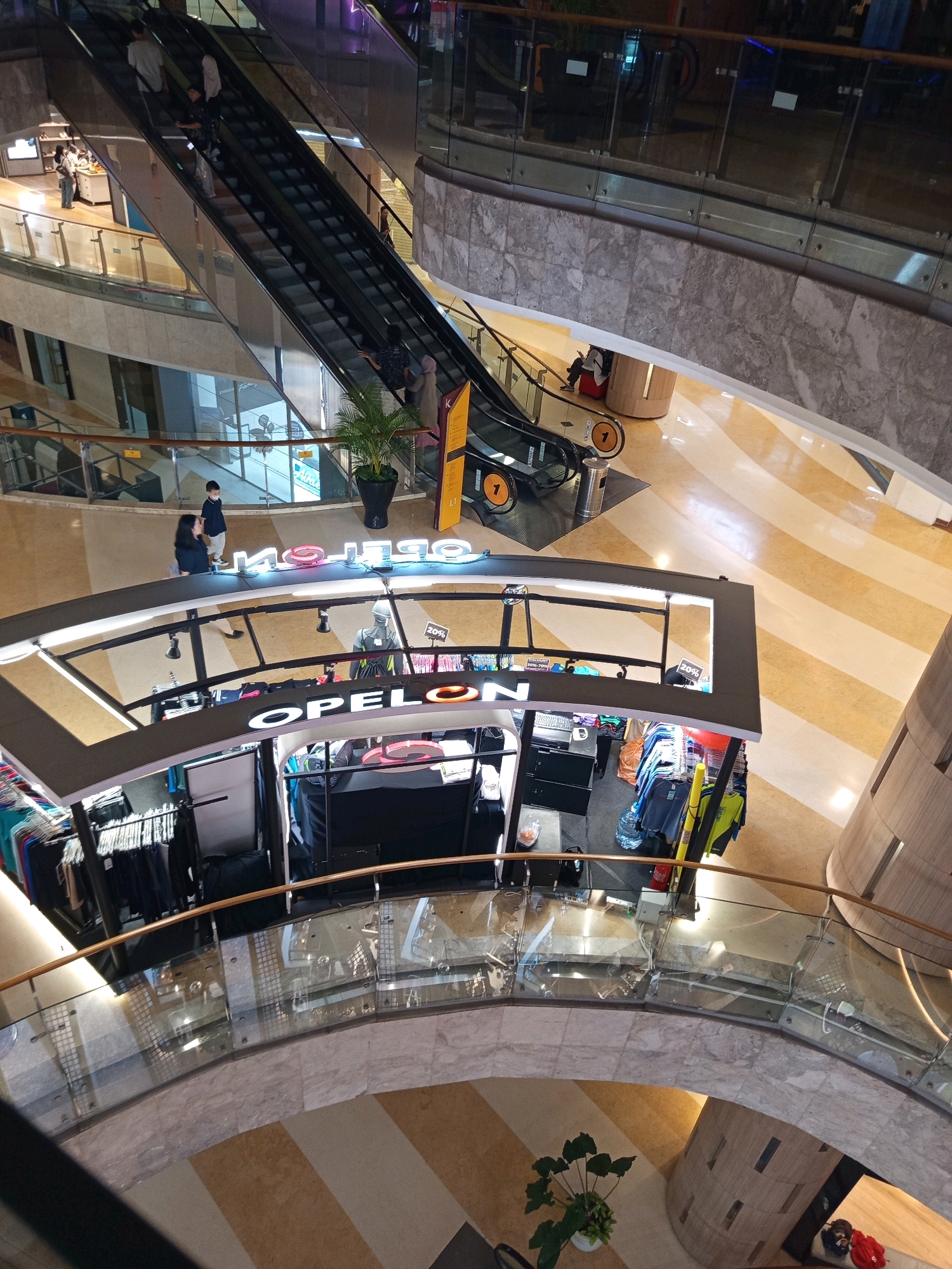 The image shows an interior view of a multi-level shopping mall in South Jakarta, Indonesia, likely taken from an upper floor looking down. In the foreground, a clothing store kiosk with the illuminated signage "OPELON" is visible. The kiosk displays clothing items, racks of garments, and a mannequin. An escalator is seen in the background, with several people using it to ascend. The mall features polished marble floors with a striped pattern, glass railings, and contemporary architecture. There are also potted plants and lighting fixtures adding to the ambiance of the commercial space.