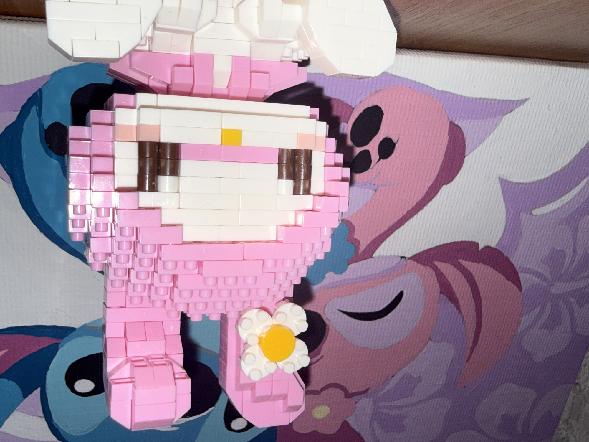 The image displays a close-up view of a pink My Melody character figurine made of building blocks. The figurine is positioned in front of a canvas painting that features stylized cartoon characters, one in pink and purple and another large blue character with a large eye. The pink character on the canvas has a paw visible. The figurine itself is detailed, with a white face, brown eyes, and a pink hood adorned with a white flower with a yellow center. The background painting appears to be a depiction of characters from the animated series Lilo & Stitch, with the pink character resembling Angel and the blue character resembling Stitch. The image is taken from a low angle, with the figurine in the foreground and the painting in the background. The surface beneath the figurine is a wooden table. There are no people or any identifiable urban elements, so it is not possible to determine the location in Rayevskiy, Russia.
