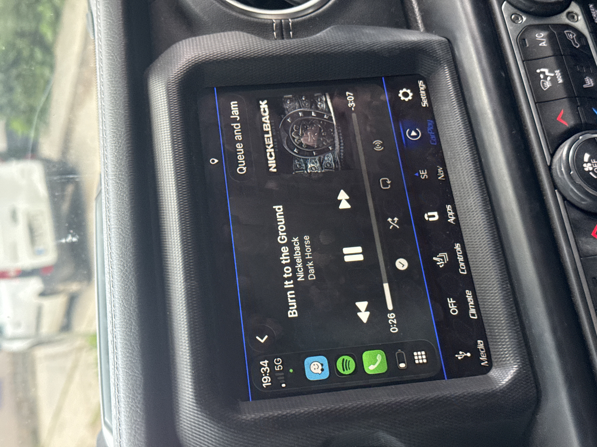 The image shows a car's infotainment screen displaying the song "Burn It to the Ground" by Nickelback from the album Dark Horse. The screen also shows other features like phone apps, navigation, climate controls, and media. Time 19:34 is indicated. It shows it is playing through the Spotify app through a possible connection to CarPlay. The location is unknown.