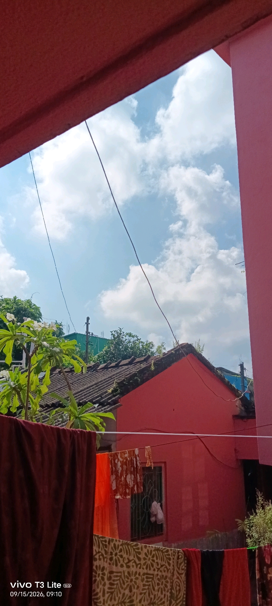 The image captures a view from a window in Baruipur, India, looking outwards towards a bright, partly cloudy sky. In the foreground, laundry hangs on a clothesline, featuring a dark red blanket, an orange garment, a patterned cloth with white floral designs, and other colorful fabrics. Beyond the laundry, a vibrant pink building dominates the lower half of the frame, with a section of its roof visible. The roof is covered with aged, grey tiles and is partially obscured by lush green foliage from a tree with white flowers. In the background, more trees and rooftops can be seen, along with electrical wires crisscrossing the sky. The lower left corner of the image displays a timestamp and device information: "vivo T3 Lite 5G 09/15/2026, 09:10".