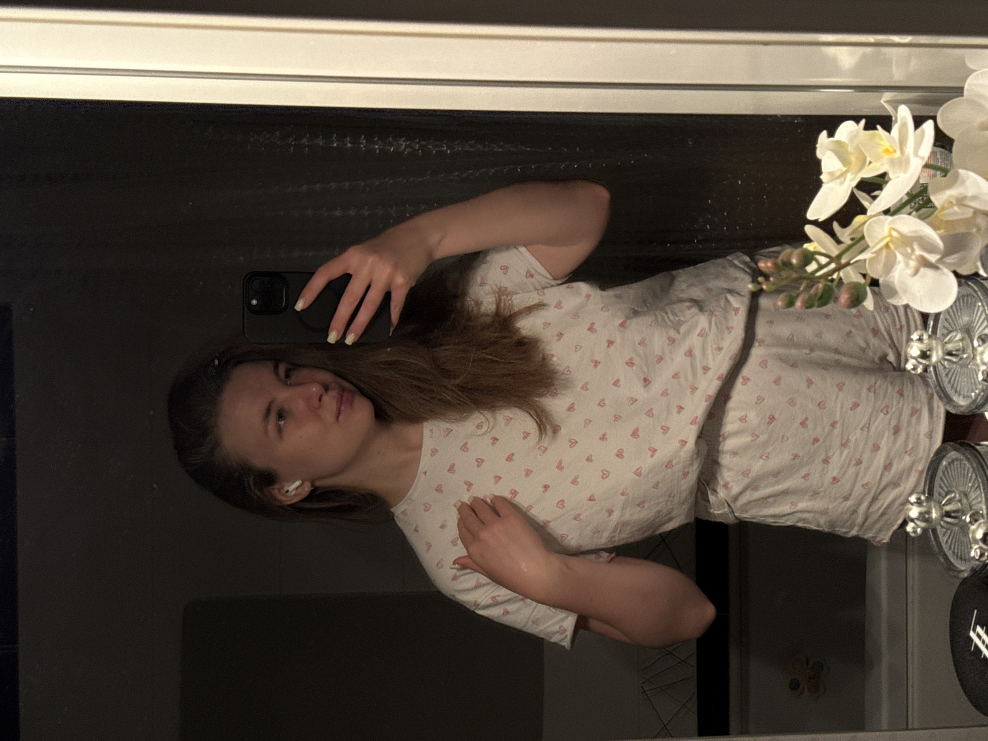 A woman is taking a mirror selfie in a room. She is wearing a white short-sleeved shirt with pink heart patterns and matching shorts. She is holding a black smartphone in her right hand. She has brown hair and is wearing an earbud in her left ear. The room appears to be a bathroom with flowers in a vase and a countertop visible. The location is not evident to be Vladimir, Russia without further context.