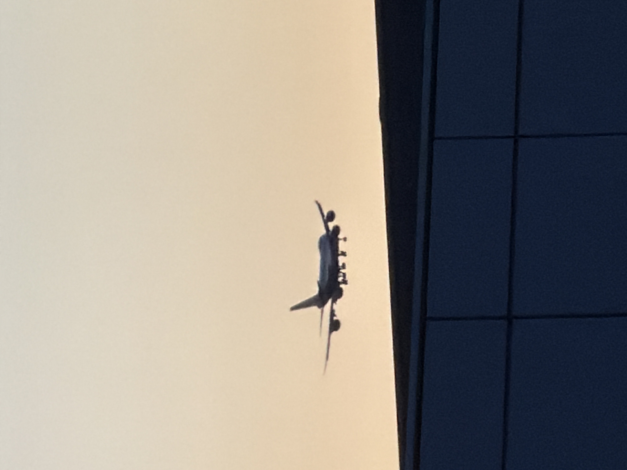 A silhouette of a large passenger airplane is captured in mid-air against a pale orange sky. The airplane, likely on its approach to land, is viewed from below and slightly to the side, revealing its wings, engines, and tail fin. In the foreground, a dark, angular structure, possibly part of a building or a bridge, is visible, with a distinctly segmented facade. The overall scene suggests an airport environment near a city. There is no specific information in the image to confirm its location in Leudelange, Luxembourg.