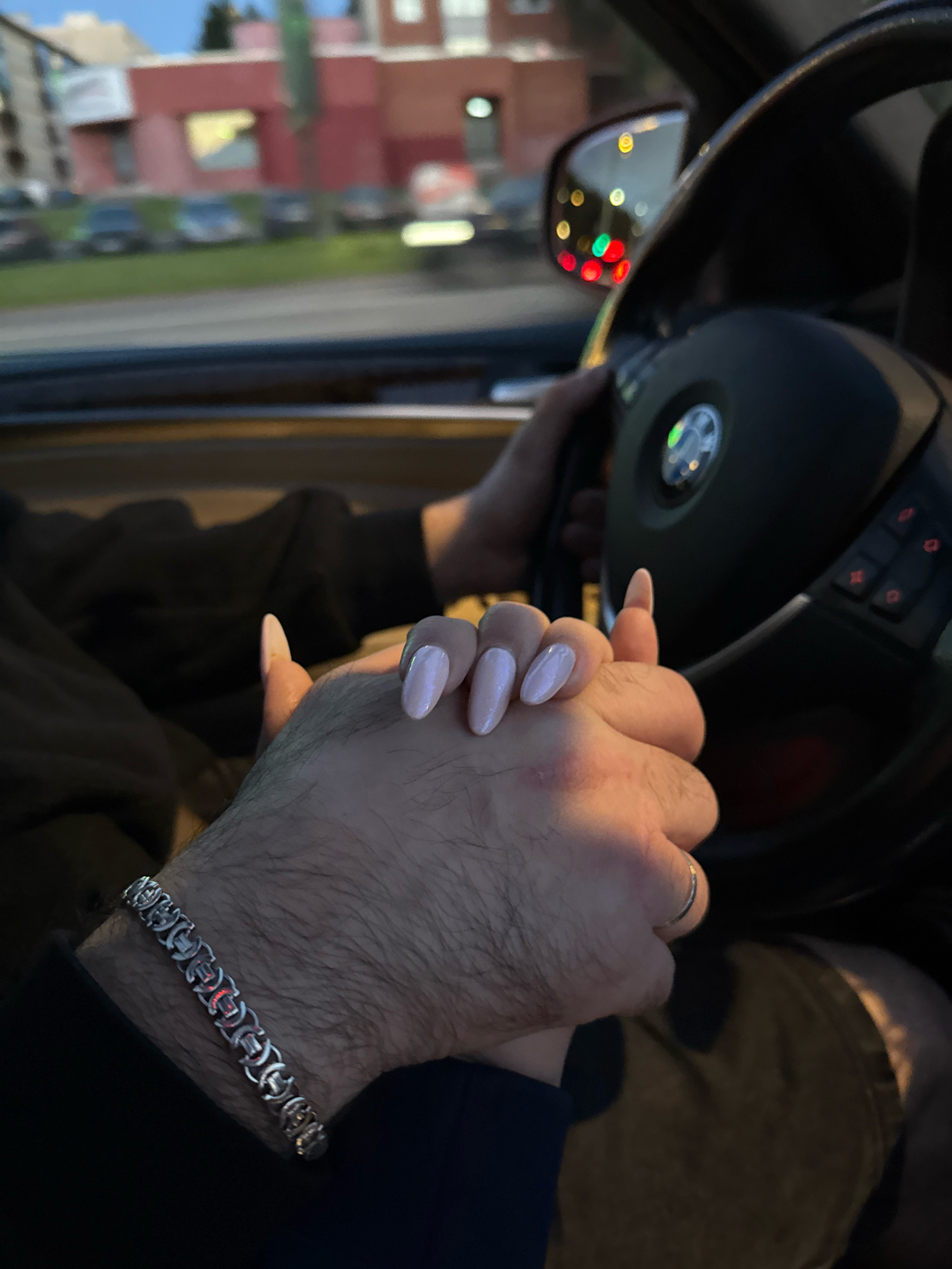 The image shows a close-up view of two people holding hands in a car. One person, presumably the driver, has their hands on the steering wheel, which is visible in the background. The other person's hands are intertwined with the driver's. The person with their hands visible in the foreground has light pink painted nails. A silver bracelet is worn on the wrist of the person whose hand is closer to the camera. A ring is visible on one of the fingers of the hand that is on top. The environment outside the car, visible through the windows, suggests an urban setting with buildings and parked cars. The lighting and colors indicate it is either dusk or dawn. No specific landmarks that identify Tallinn, Estonia are visible.