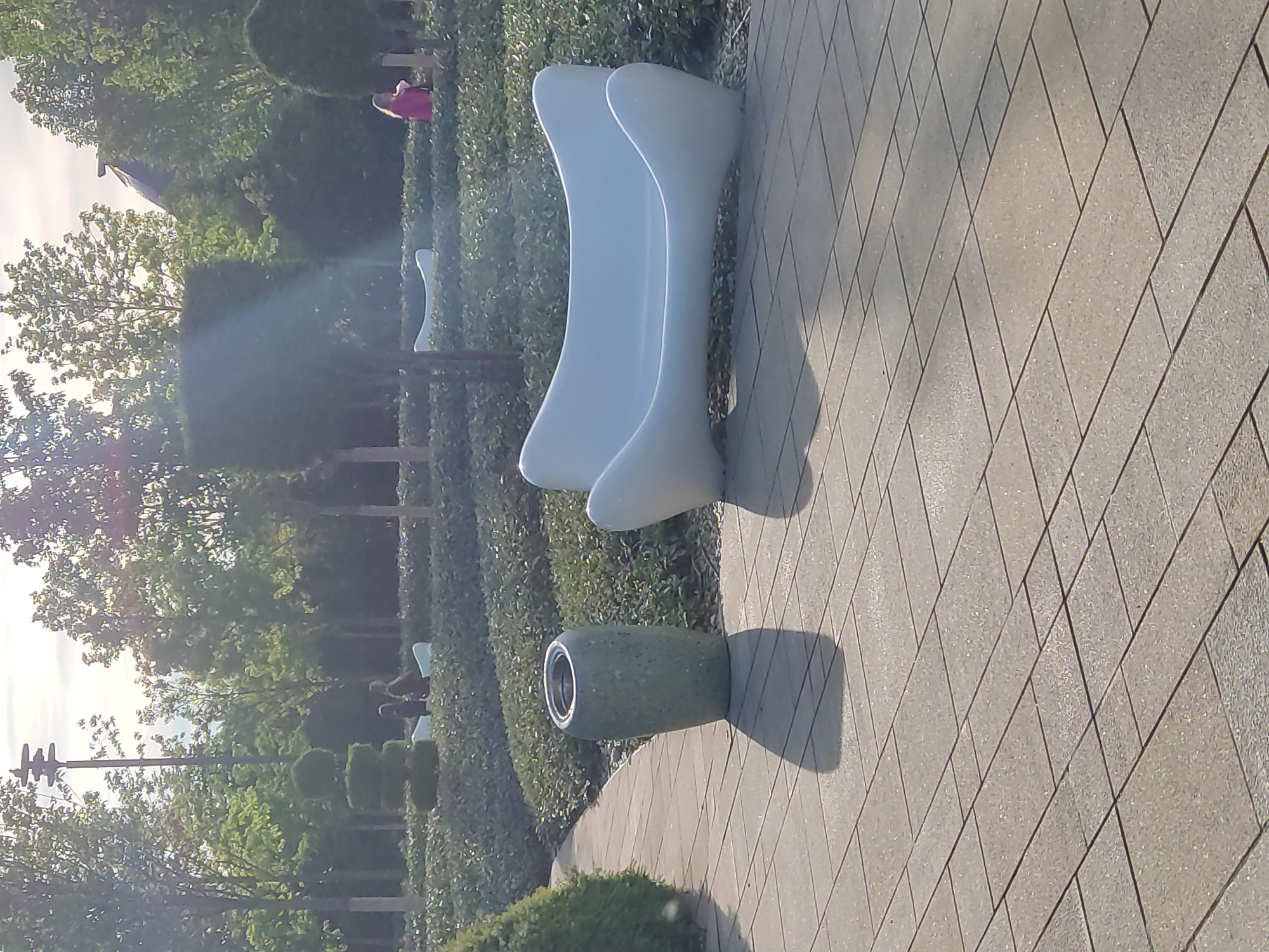 Here is a description of the image:
The image shows a park setting, likely in Krasnodar, Russia. Two modern-style white benches are situated on a paved walkway made of light gray, diamond-patterned pavers. A cylindrical trash receptacle is positioned near the benches. The background consists of manicured hedges, trees, and some distant structures, including what may be a building with a dark-colored roof. A person wearing a pink shirt is visible standing in the distance near the hedges. The light is bright, creating shadows that suggest the time of day is around noon.