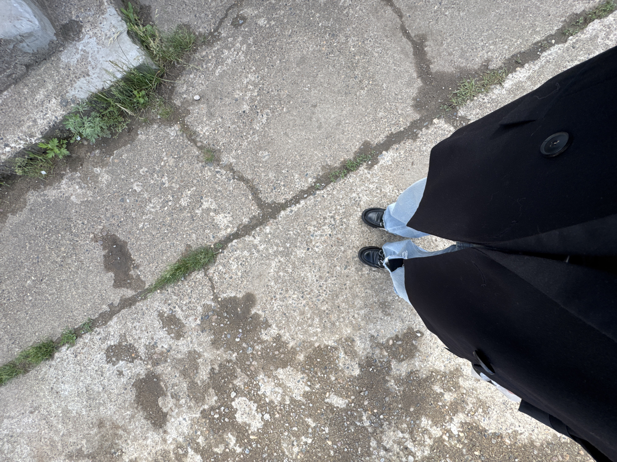 The image, taken in Chaikovsky, Russia, offers a top-down perspective of a person standing on a concrete surface. The lower parts of two legs are visible, dressed in light blue jeans and black shoes, possibly loafers, which feature a chain-like embellishment across the vamp. A black garment, likely a long coat or dress, extends into the frame from the right, covering parts of the legs and featuring a single large black button.
The ground consists of grey, speckled concrete, heavily cracked throughout. Green grass and various weeds protrude from these cracks, suggesting an unkempt or naturalized outdoor area, such as a sidewalk or pathway. Scattered across the concrete are irregular dark patches, indicating wetness, possibly from recent rain or spilled liquid. The lighting is bright and natural, consistent with daytime. There is no discernible activity beyond standing. No text is visible within the frame.