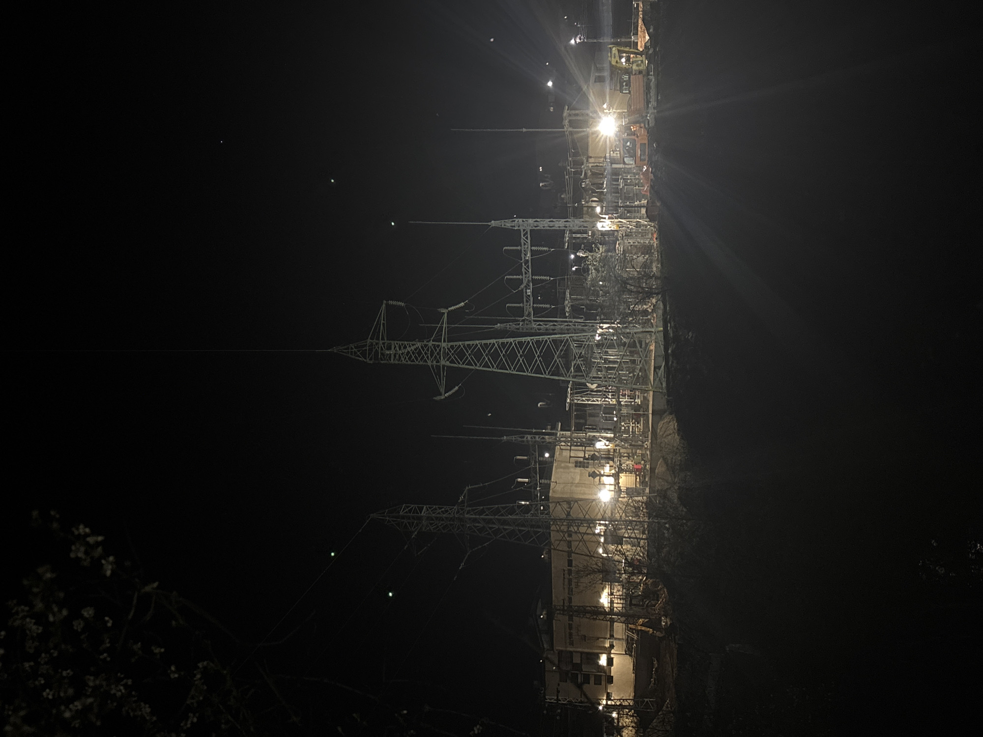 The image depicts an electrical substation at night. It features high-voltage power lines suspended from steel towers, along with various electrical components, transformers, and switchgear. Some buildings and vehicles can be seen inside the substation perimeter. The location is likely near Zlatograd, Bulgaria. There are no people visible. The surrounding environment is dark, with some vegetation visible in the foreground.