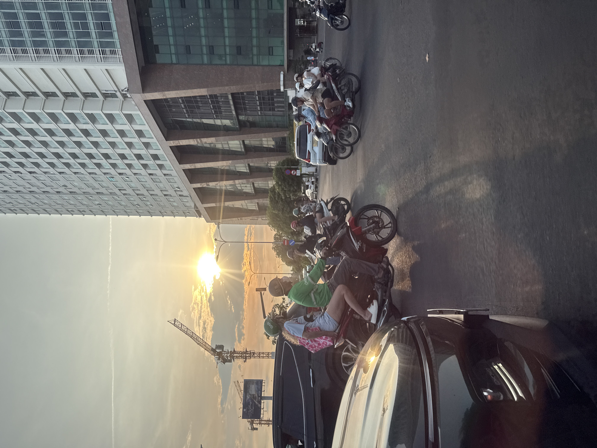 In Nha Trang Ward, Vietnam, a street scene unfolds at sunset. A modern, tall building with multiple floors and glass balconies dominates the background, its facade reflecting the warm, orange glow of the setting sun. In the foreground, several motorcycles navigate the busy street, many carrying multiple passengers. One motorcycle in the center has a woman in a light blue top and shorts, wearing a green helmet, riding as a passenger, with a pink patterned backpack on her shoulders. She is sitting behind a man wearing a green shirt and a beige helmet, who is operating the motorcycle. The street is asphalt, with visible lane markings. A large crane, indicative of ongoing construction, is visible against the sky, which is filled with streaks of clouds and the bright sun. Several cars are also present, including a dark SUV in the foreground with its front end angled towards the viewer, its headlights subtly illuminated. The overall atmosphere suggests a typical evening commute in a bustling urban environment.