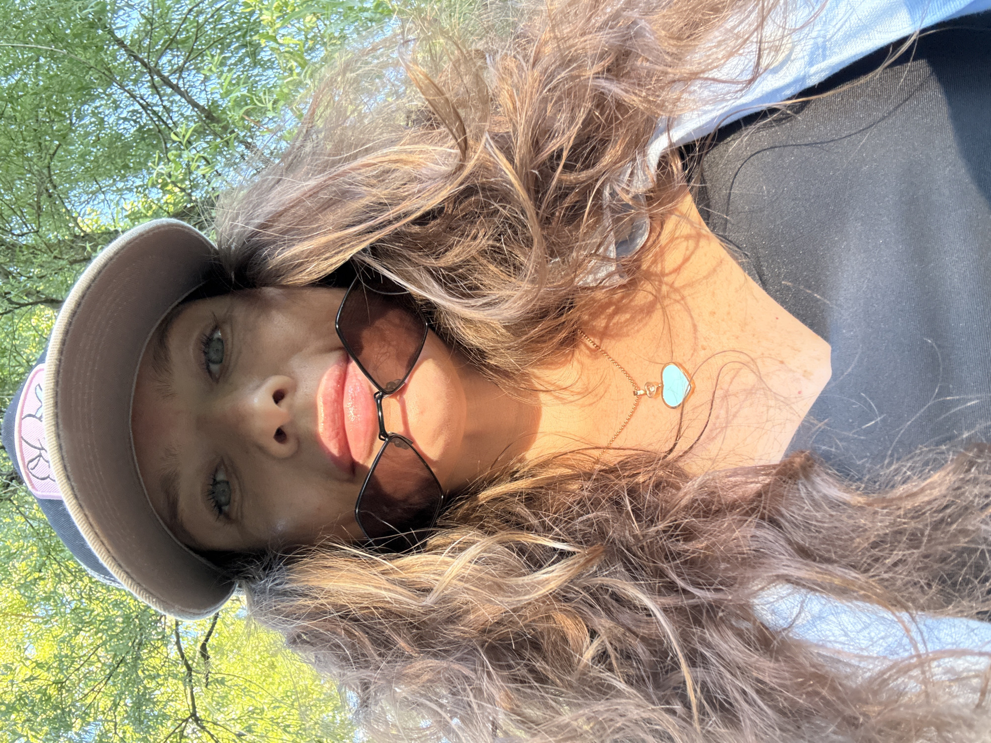 This media file captures a close-up, low-angle selfie of a young woman outdoors. The subject is wearing a baseball cap, sunglasses, and a necklace with a heart-shaped pendant. Her long, wavy brown hair is prominent in the frame, partially obscuring her face. The background consists of blurred green foliage and blue sky, suggesting a sunny day in a park or wooded area. The lighting indicates it is daytime, likely late afternoon due to the warm, golden quality of the sunlight illuminating her skin and hair. The overall mood is casual and relaxed. There is no discernible text in the image. The location is specified as Moscow, Russia.