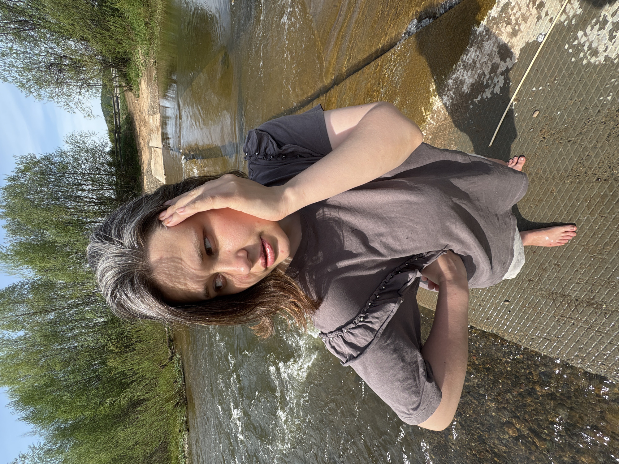 A woman stands on a concrete structure beside a river in Усть-Тунтор, Russia. She wears a gray t-shirt with ruffled sleeves and light-colored shorts. Her hair is brown with gray streaks. She holds her hand to her forehead and looks toward the river. The river flows from left to right in the background, surrounded by trees. The sky is visible at the top left corner.