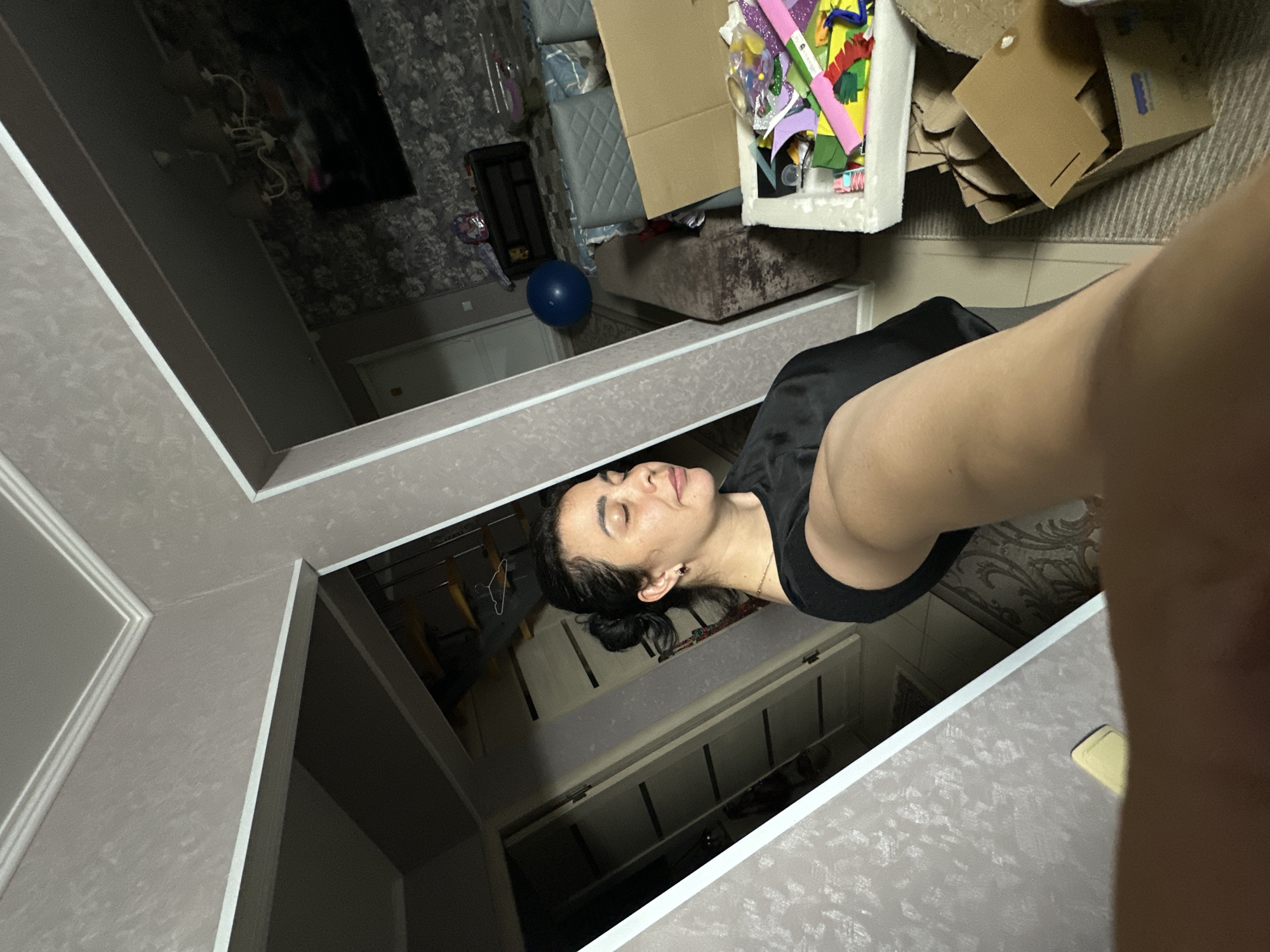 A mirror selfie captures a woman with dark hair styled in a low bun. She wears a black top and is positioned facing upwards in the mirror's reflection. The reflection reveals a room cluttered with items, including boxes filled with colorful craft materials and a blue balloon. The room's décor includes patterned wallpaper. A clothing rack can be seen in the background of the reflection. The image appears to be taken indoors. The city and specific location are unknown.