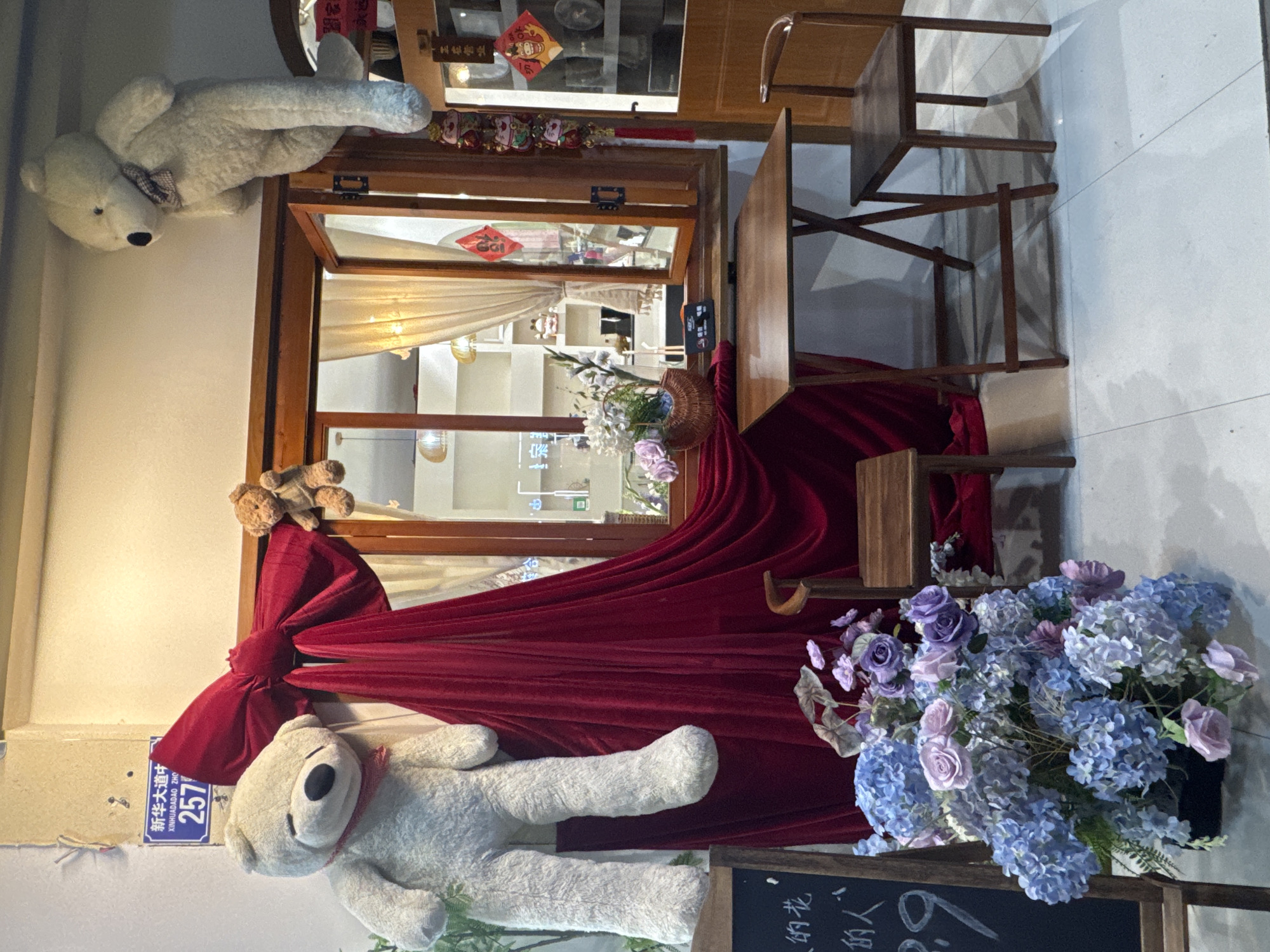 Here is a description of the image:
The image captures a whimsical, ornate storefront display, likely in Qianjiang, China. A large wooden-framed window acts as a stage, adorned with a thick, red velvet drape tied into a large bow on the left side. Two plush, white teddy bears add to the playful scene: one seemingly suspended above the window frame, the other draped at the bottom. A basket of flowers sits within the window frame, contributing to the overall decorative effect.
To the right of the window, a simple wooden table and chair are arranged, suggesting a small seating area or display setup. A small chalkboard stands in front of the flower basket. A blue street sign on the left side of the frame reads "Xinhua Dadao Zhong 257" indicating the location is on Central Xinhua Avenue. Chinese characters are also present on the display, giving more clues about the setting. The overall aesthetic leans towards a charming, themed decor, potentially for a restaurant or boutique.