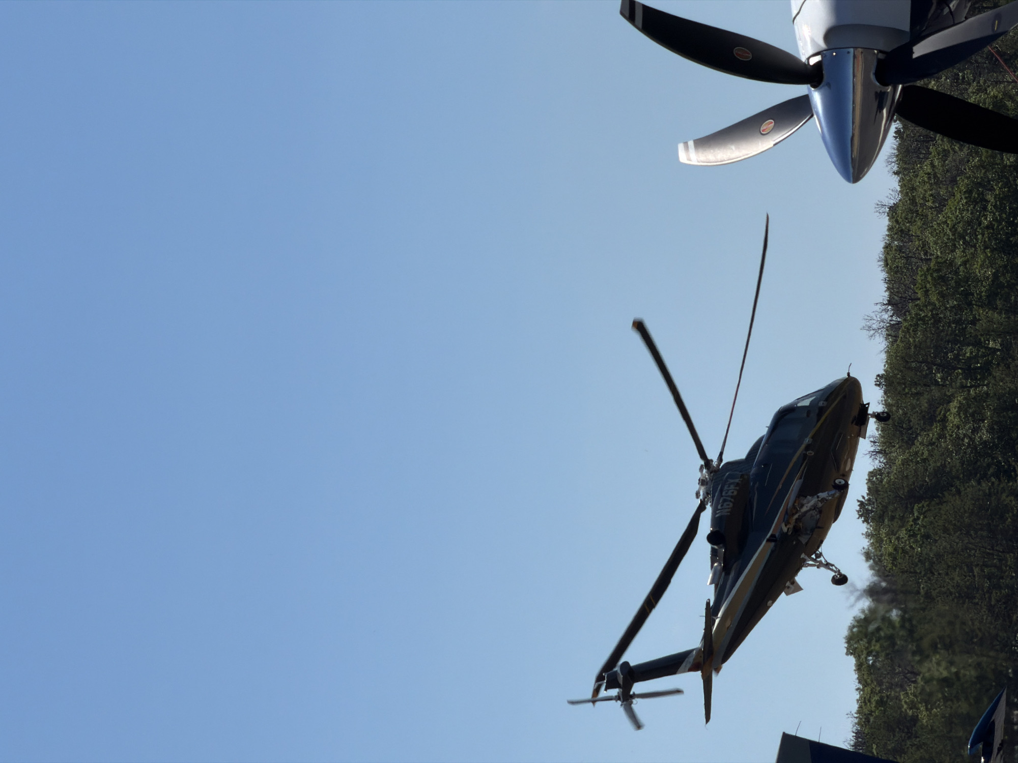 A dark-colored helicopter with registration number N676FX is captured in mid-air, appearing to be either taking off or landing. The helicopter has a sleek design, with gold accents along its body. Its landing gear is extended, suggesting an imminent touchdown or recent lift-off. In the foreground, the prominent propeller of another aircraft, likely a turboprop plane, is visible on the right side of the frame, its blades sharp and metallic. The background features a dense canopy of green trees under a clear, bright blue sky, indicative of a daytime scene. The overall impression is one of aviation activity, possibly at an airport or a private airstrip in a lush, wooded area. The location is specified as the Town of East Hampton, United States of America.