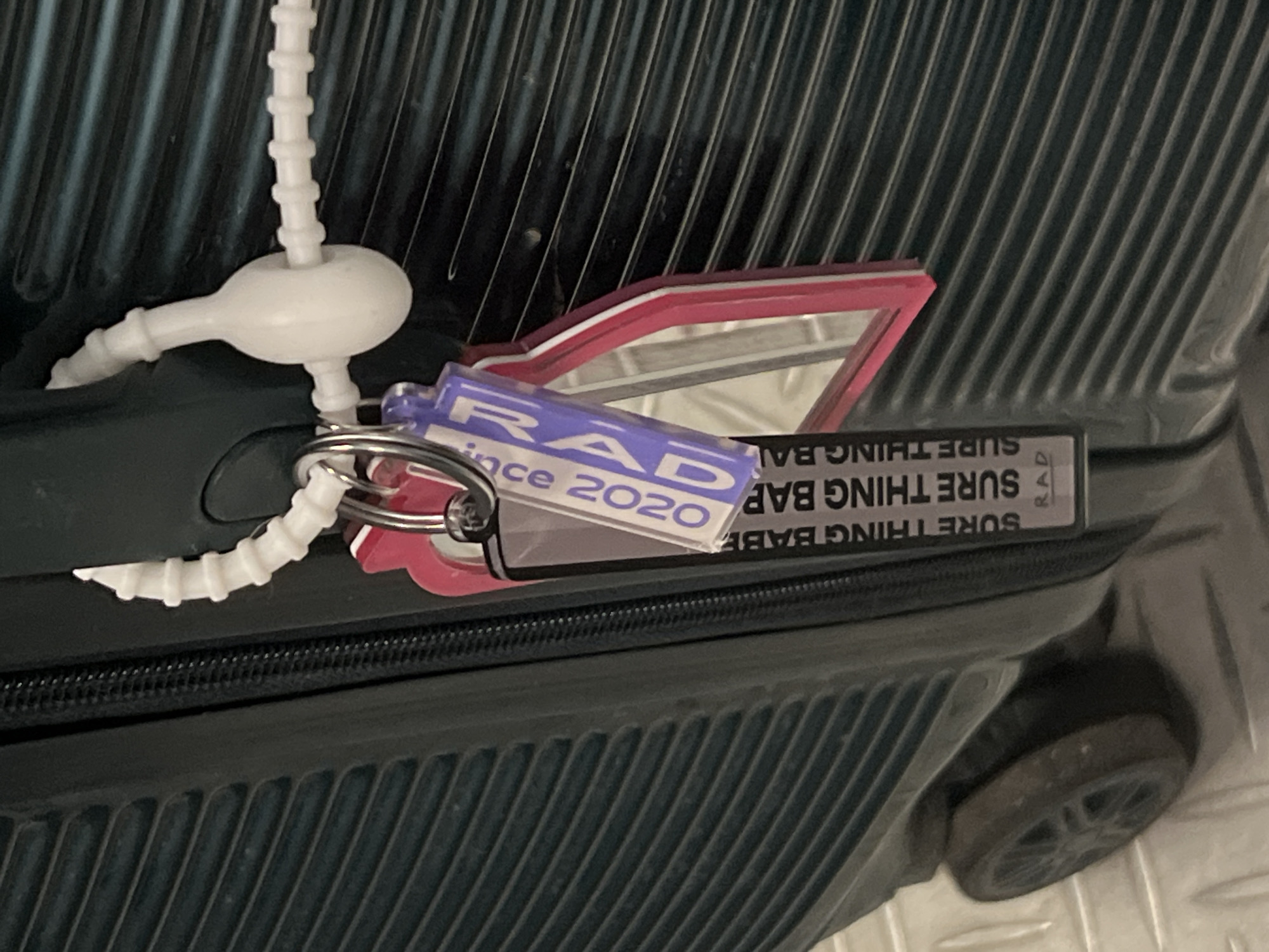 A close-up shot shows a section of a dark-colored suitcase, possibly black or dark teal. The suitcase has a ridged design on its surface. Attached to the zipper pull are several keychains, including one that says "RAD since 2020," another shaped like a car, and a white plastic tag. The suitcase appears to be resting on a light-colored, possibly tile floor. A wheel of the suitcase is visible in the lower right corner. There is no indication of Moscow, Russia, or any people in the image.