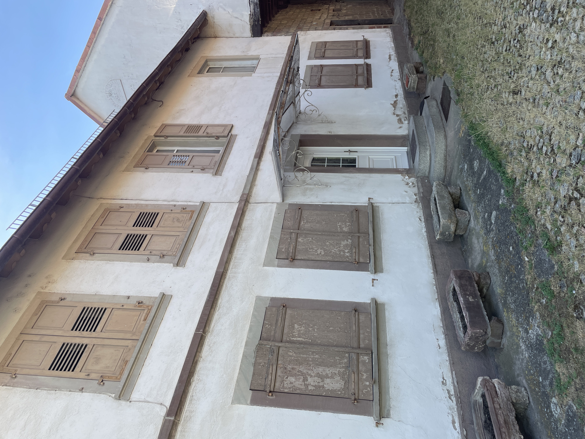 The image shows the exterior of a historic building with white stucco walls and wooden shutters. The building has multiple stories with several windows. Some windows have louvered shutters that are closed, while others have open shutters revealing the glass behind them. The architectural style suggests an older building, possibly in a European village. The ground next to the building is covered with grass and cobblestones, and there are some stone troughs or planters visible. The sky is a clear blue, indicating a sunny day. The sunlight casts shadows on the building, suggesting it is daytime, likely late morning or early afternoon. There is no visible text in the image. The overall impression is of a peaceful, historic setting in Gerstheim, France.
