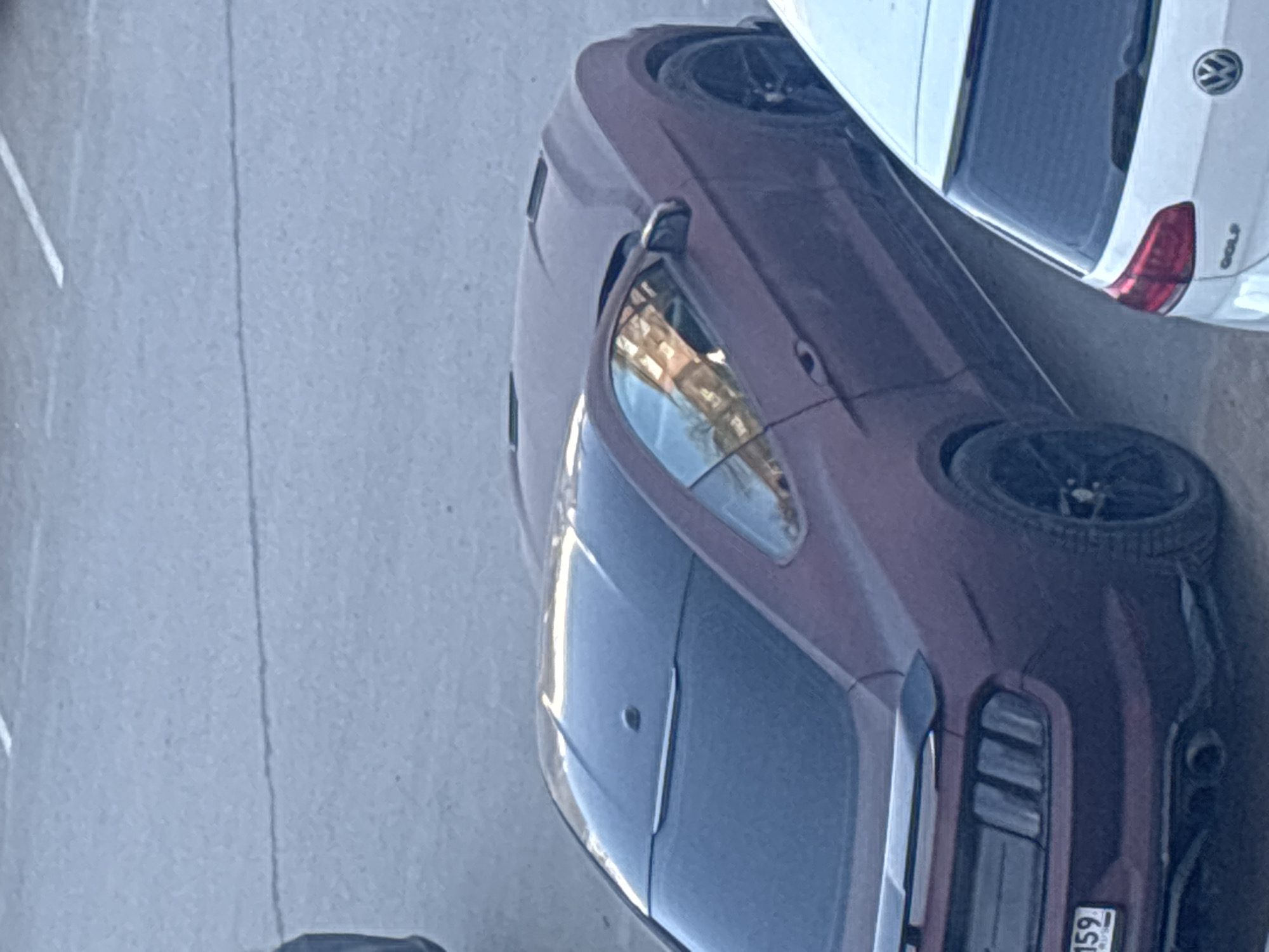 The image shows two cars parked in an urban environment, possibly in Perm, Russia. One car is a maroon/burgundy color, and the other is white. The white car is partially obscured, but visible from the rear, and appears to be a Volkswagen Golf based on the emblem. The maroon car is positioned next to the white car, its front end closer to the camera. The background shows a paved area, likely a parking lot or street. The maroon car has a license plate visible.