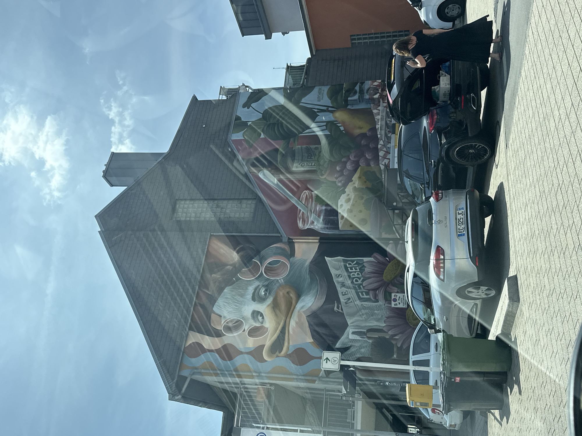 This image shows a mural on the side of a building in Käerjeng, Luxembourg. The mural depicts a duck-like creature with curlers in its hair, reading a newspaper titled "NEWS FERBER". To the right of the duck, the mural continues with an illustration of various food items, including grapes, cheese, and a jar of jam. In the foreground, several cars are parked. A woman in a black dress is standing next to an open black SUV, appearing to place items into the trunk. Two trash bins are visible on the left side of the image, and a yellow mailbox is positioned behind them. The scene is set on a paved area under a bright, partly cloudy sky.