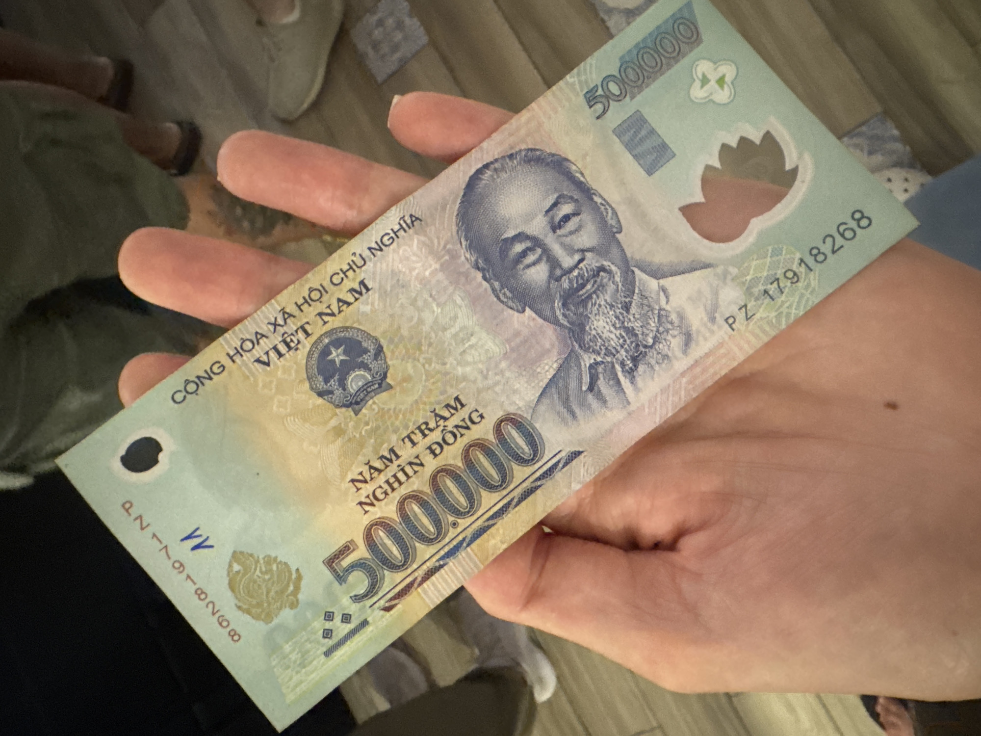 A close-up shot shows a hand holding a Vietnamese 500,000 dong banknote. The banknote features a portrait of Ho Chi Minh, the national emblem of Vietnam, and the text "VIỆT NAM" and "500.000". The banknote is light green and yellow. The background is blurred, but appears to be a tiled floor. The lighting suggests daytime. The hands are light-skinned. The activity appears to be showing or displaying the banknote.