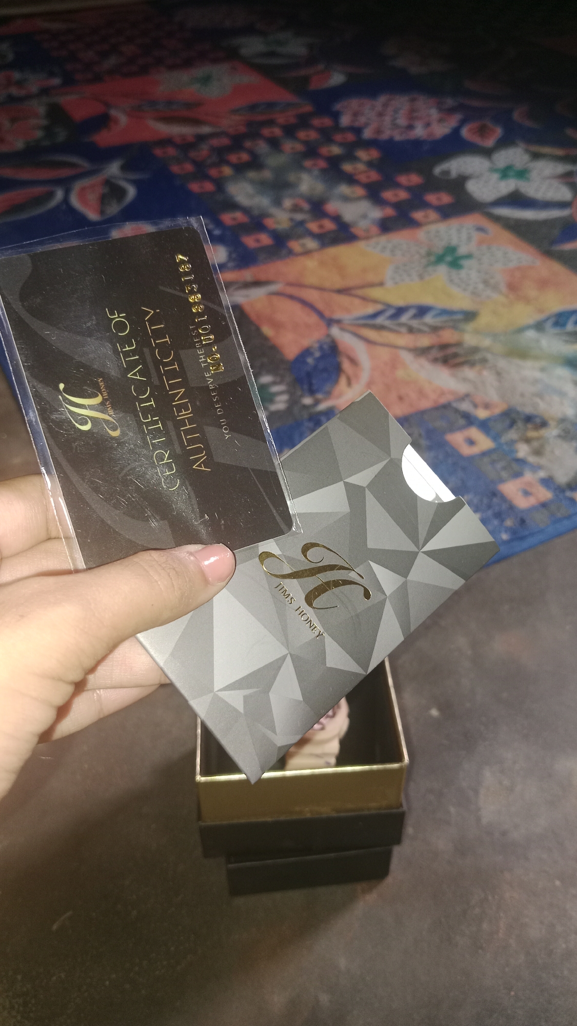 In Wono Sari, Indonesia, a hand is holding a "Certificate of Authenticity" card and a Jims Honey card. The certificate is black with gold lettering and a gold "H" logo. The Jims Honey card is gray with a geometric pattern and a gold "H" logo, also reading "JIMS HONEY". The cards are positioned over an open black and gold box. The background is a colorful patterned fabric.
