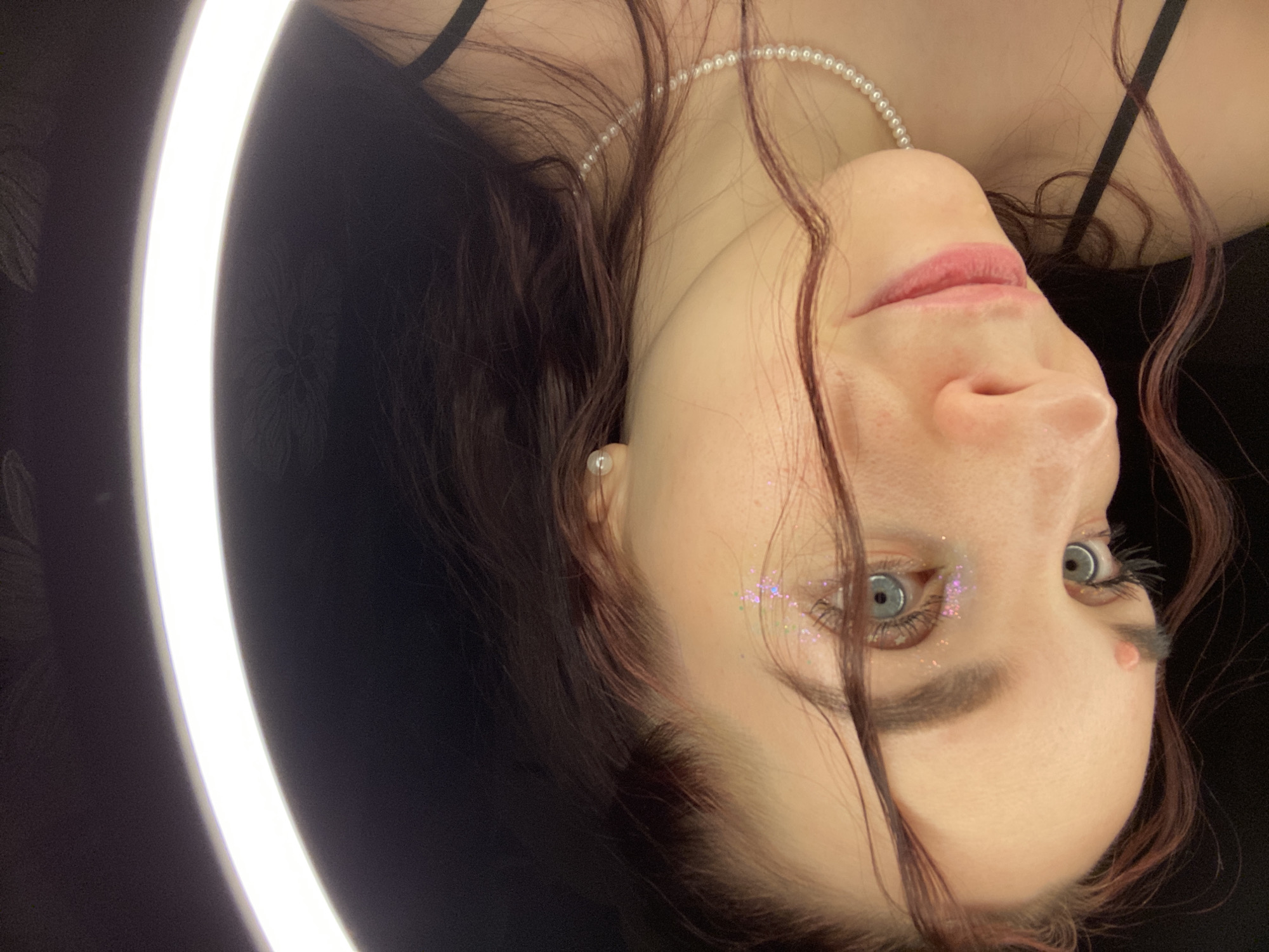 A woman is shown from the chest up, turned 90 degrees to the left. She has light skin, light blue eyes, dark eyebrows, and light pink lips. She is wearing a pearl necklace and a stud earring in her left ear. There is glitter on the right side of her face, below her eye. Her dark hair is visible. She is wearing a black top with thin straps. A light ring is visible on the left side of the photo. The location is unknown, as there is no evidence of the city of Chaikovsky, Russia, in the image.