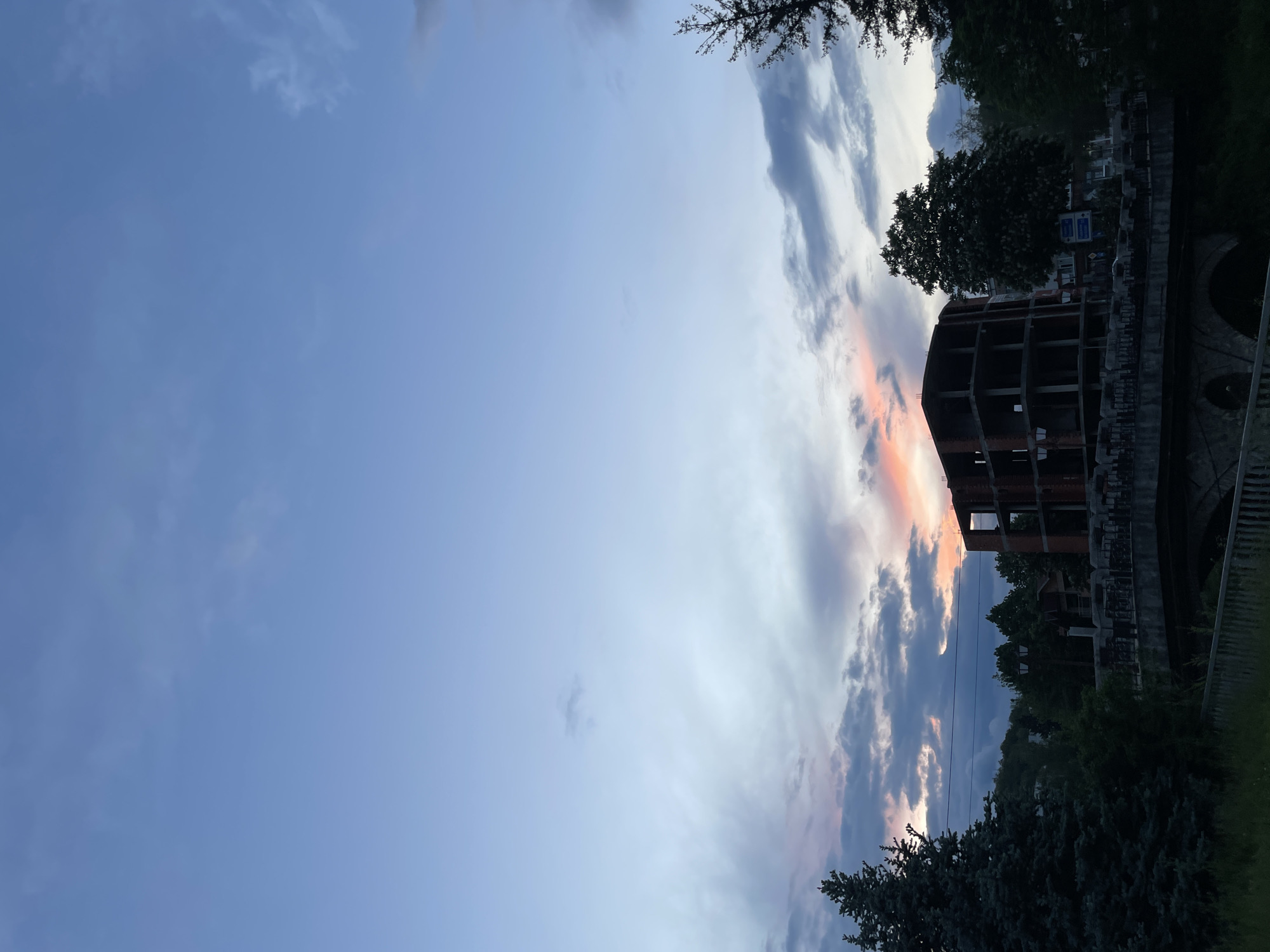 The image shows an unfinished building with a concrete frame in Zlatograd, Bulgaria. The structure is located next to a bridge or retaining wall, with trees partially obscuring the view. The sky is mostly blue with clouds tinged with orange from the setting sun. The scene appears to be taken in the evening.