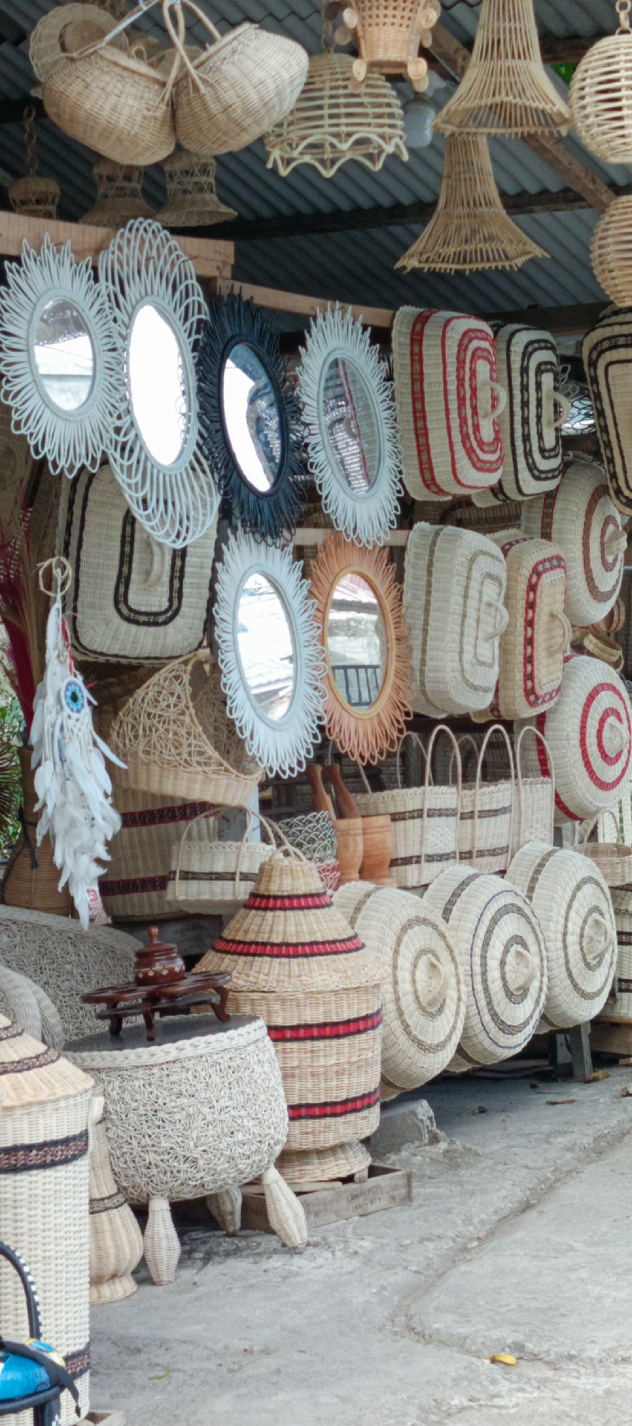 The image depicts a stall selling woven handicraft items in Tanjong, Indonesia. The stall is filled with a variety of products made from rattan and other natural fibers. Prominently displayed are woven baskets of different sizes and shapes, some with handles, and cylindrical containers with red and black striped patterns. Several oval-shaped mirrors with decorative, sunburst-like woven frames are hung on the wall. Hanging from the ceiling are woven lampshades and baskets. In the foreground, a circular woven table with a small wooden container sits on top of a large, round woven object supported by four legs. The stall is set up outdoors, with a corrugated metal roof visible above the display. The ground is made of rough concrete. A small white decorative item with blue beads and white feathers hangs from a string on the left side of the stall. The scene is brightly lit, suggesting daytime.