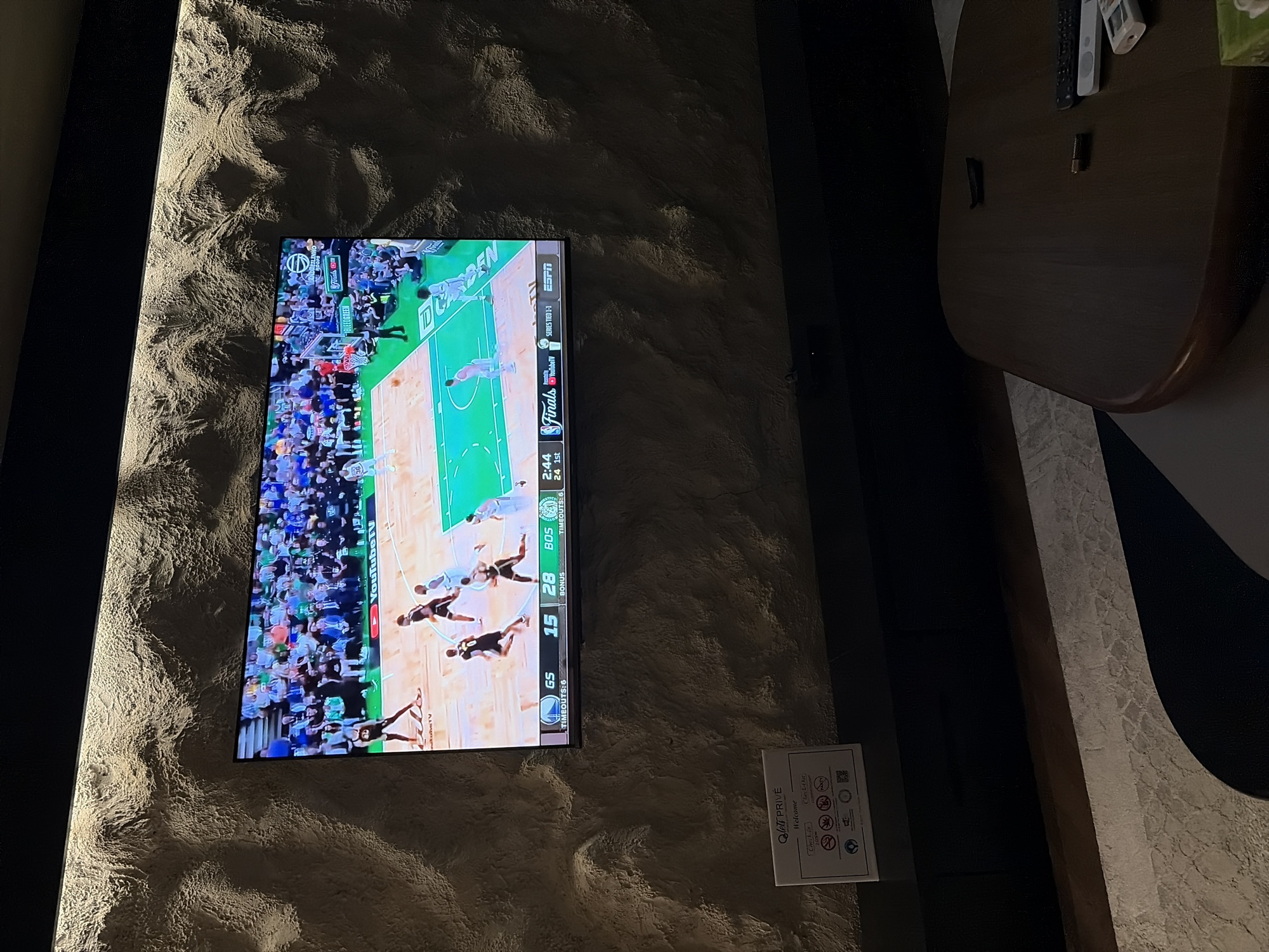 The image captures a dimly lit interior space, likely a private room or suite in a hotel within Abuja, Nigeria. A large flat-screen television is centrally mounted on a wall featuring a dramatic, textured finish, possibly stucco or plaster. Accent lighting from above highlights the uneven surface, casting strong shadows and lending a sophisticated, cozy atmosphere.
The television displays a professional basketball game, identified as the "Finals." The broadcast is from "ESPN" and streamed via "YouTube TV." The visible scoreboard indicates "GS 15" and "BOS 28" with "2:44" remaining in the "1st" quarter. Game statistics also show "TIMEOUTS: 6" for both teams and a "BONUS" indicator. Notable on-screen text includes "GREENBRIER" and "CHASE" (likely sponsors), and player information showing "CURRY" next to a player number (partially obscured). Players in dark and light jerseys are seen actively engaged in the game.
To the right, a dark, polished wooden table with a curved edge holds two remote controls and a small, dark cylindrical object. Below the television, a white informational sign is affixed to the wall. This sign clearly displays "Qlett PRIVÉ" at the top, followed by "Welcome," "Check-in," and "Check-Out." It also features a QR code and a series of universal iconography, such as symbols for "no smoking," "no pets," and potentially others related to room policies. The overall setting suggests an evening scene, designed for relaxation and entertainment.