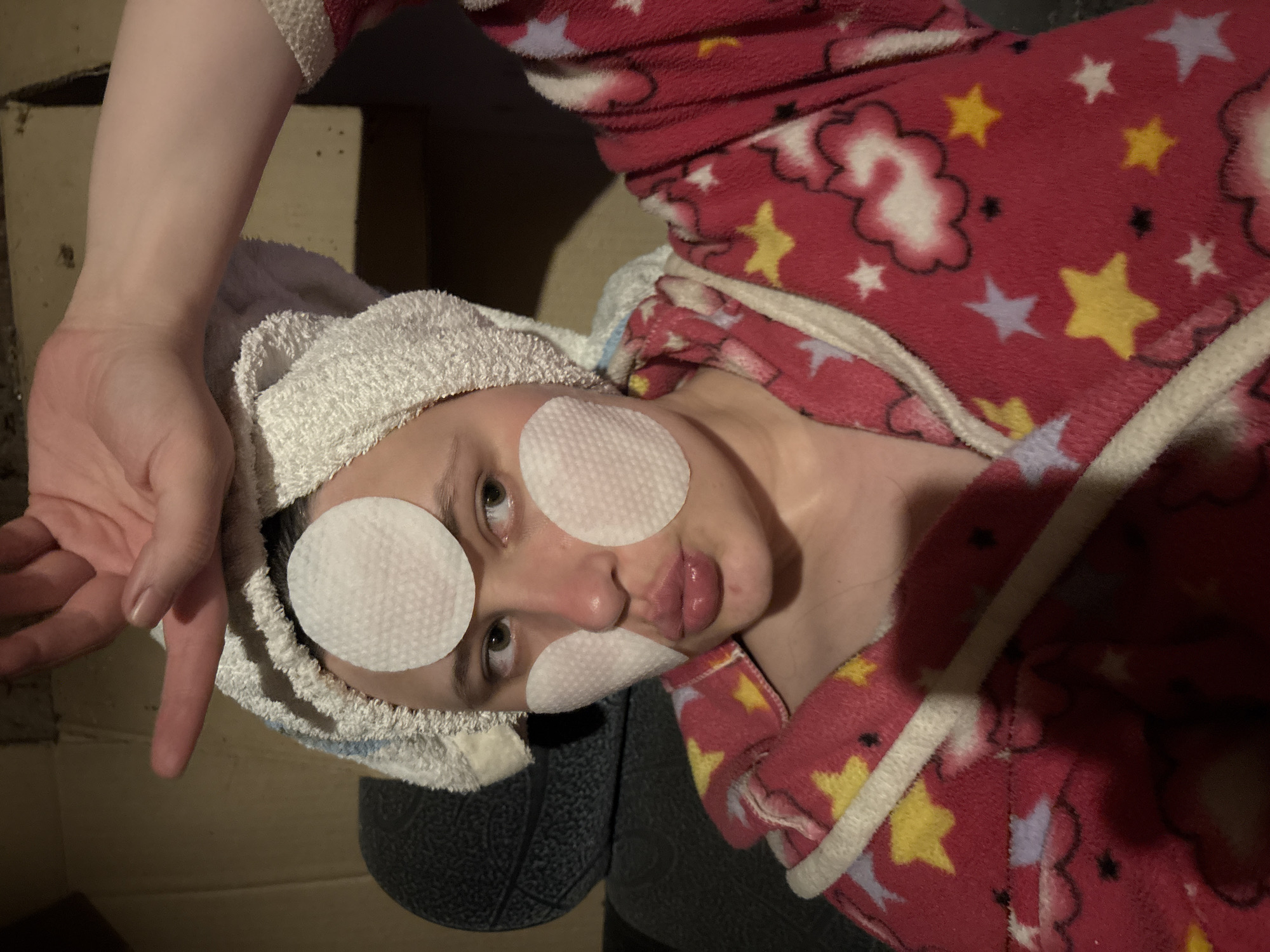 A person is lying on what appears to be a cushioned surface, possibly a chair or sofa. The person is wearing a white towel wrapped around their head and has white cotton pads on their forehead, cheek, and nose. The person is wearing a red robe or pajamas with a pattern of stars and clouds. An arm and hand are visible in the upper left corner of the image, with fingers extended. The background appears to be a cardboard box.
Given the available information, it is not possible to determine if the location is in Nizhnyaya Chermoda, Russia. This would require additional information or context.