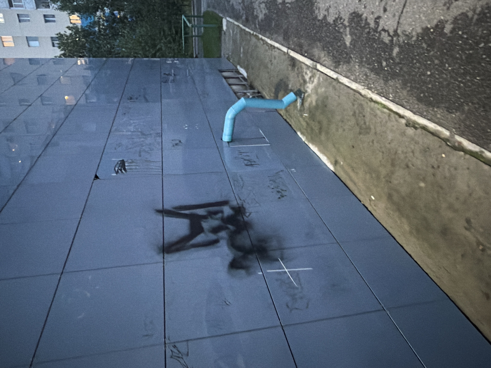 The image depicts the exterior of a modern building, possibly a residential or commercial structure, in Surgut, Russia. The building's facade is made of large, dark gray, rectangular panels, arranged in horizontal rows. There are visible horizontal seams between these panels.
Graffiti is present on the facade. In the center-left of the image, a prominent black spray-painted design, possibly stylized letters or a symbol, is visible. To the left of this, a white cross symbol is drawn. Other faint markings and smudges are also present on the wall.
A light blue, curved drainpipe is attached to the building on the right side, extending outwards and downwards towards the ground. The pipe appears to be made of plastic. Near the base of the pipe, on the ground, there are some indistinct objects, possibly debris or discarded items.
The ground surface in front of the building consists of a concrete sidewalk or walkway adjacent to a darker, possibly asphalt, surface. The concrete area appears wet and slightly discolored, with some patches of green moss or algae visible along the edge.
In the background, to the right of the building, glimpses of other buildings and trees can be seen. The building in the background appears to be a multi-story apartment complex with illuminated windows. The overall lighting suggests it is either early morning or late evening, with a somewhat overcast sky. There are no people or animals visible in the frame.
