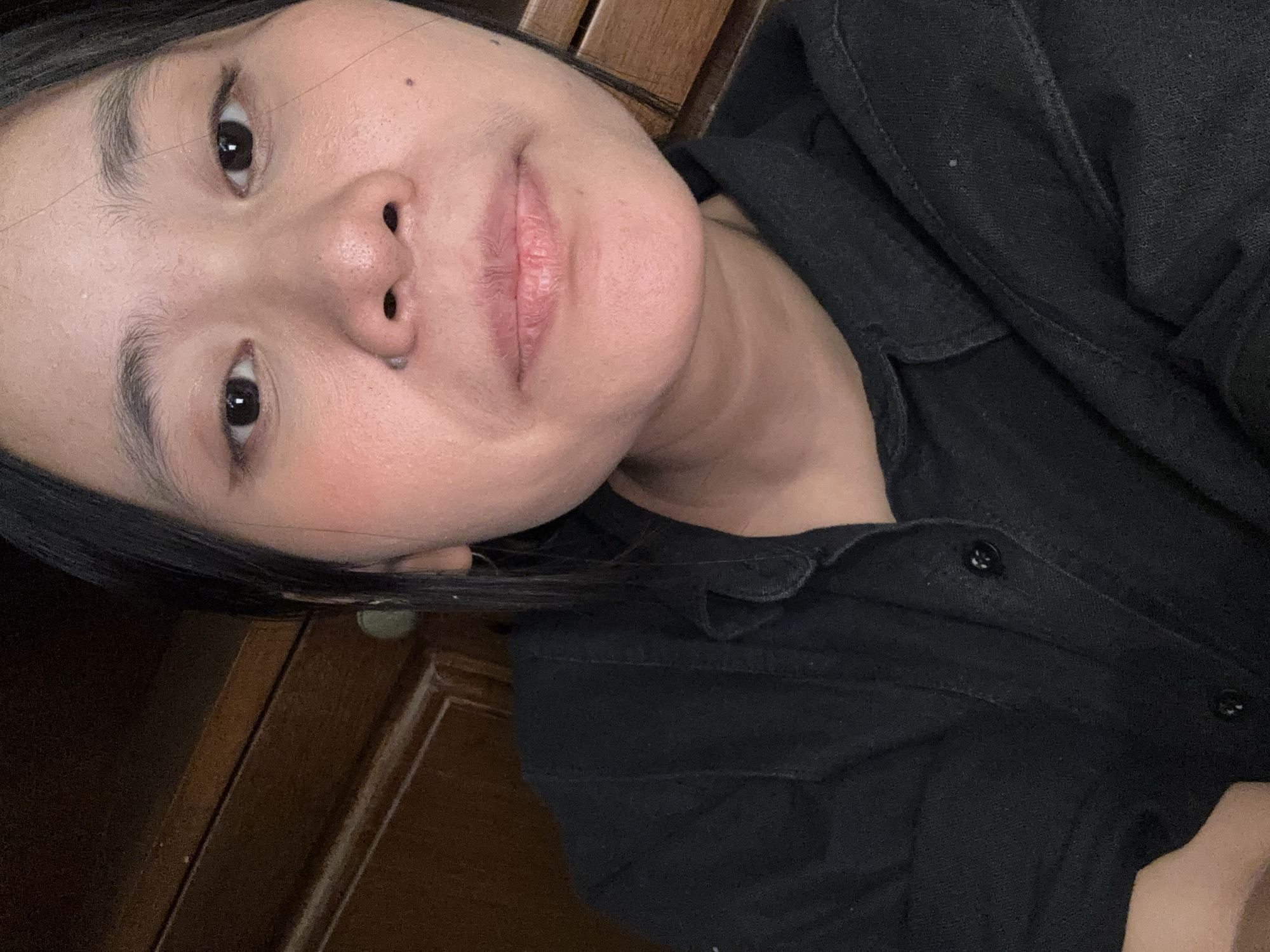 The image shows a close-up, selfie-style shot of a young East Asian woman. She has dark hair, parted in the middle and framing her face. Her eyes are dark and open, looking directly at the camera. She is wearing a black collared shirt. The background appears to be a wooden cabinet with horizontal panels. The lighting is somewhat dim, with the primary light source coming from the front, illuminating her face. The overall impression is that of an indoor setting. There are no explicit indicators of a specific location within South Jakarta, Indonesia, beyond the implicit possibility that the individual resides there.