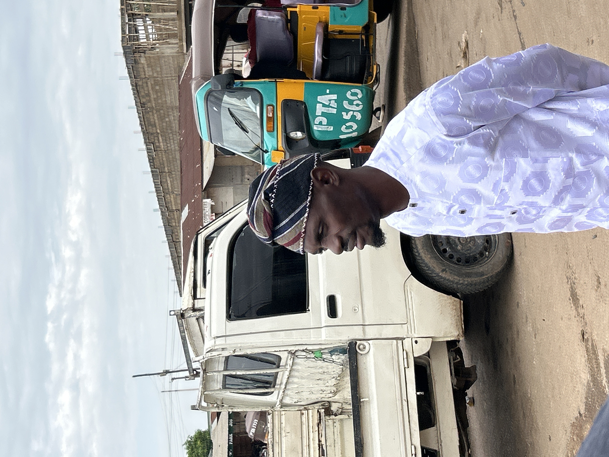 A man wearing a white patterned garment and a striped cap stands in a street scene in Jos, Nigeria. He is looking to his left, with a slight smile. Behind him, a white truck with a dented bed is parked, and to his right, a teal and yellow motorized tricycle, also known as a "Keke Napep," is visible. The ground is dirt and pavement. In the background, there are buildings under construction and some other structures. The sky is cloudy. The man's attire and the presence of the Keke Napep suggest a typical urban environment in Nigeria.