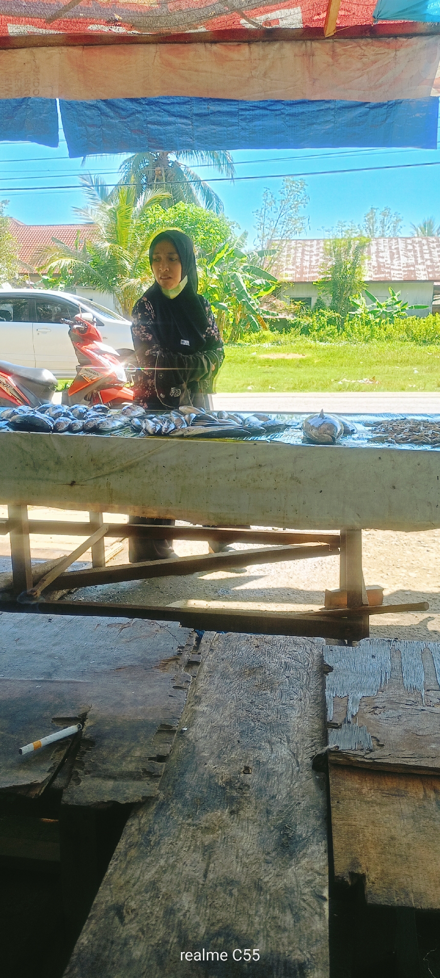 A young woman wearing a black hijab and a patterned dress stands behind a table laden with fresh fish. She is looking down and to her right, her hands clasped in front of her. The table is covered with a white tarp, and a variety of fish, some whole and some filleted, are arranged on it. Behind the table, a red motorcycle is parked next to a white car. The scene appears to be an outdoor market under a makeshift shelter made of blue and beige tarps. In the background, tropical foliage and houses with red roofs are visible under a bright blue sky. The foreground shows weathered wooden planks, with a lit cigarette resting on one of them. The overall setting suggests a street-side fish stall in a tropical urban environment.