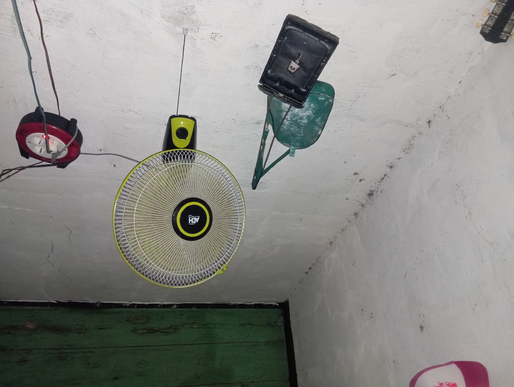 In a dimly lit, sparsely furnished room, likely in Rejosari, Indonesia, a wall-mounted electric fan with a yellow grill and black and yellow base is the central focus. The fan is plugged into a red and black extension cord reel, which is also mounted on the wall. Wires snake from the fan and the extension cord, connecting to an unseen power source.
To the left of the fan, two bags hang from the wall. A green, worn shoulder bag with a darker green bottom is suspended by a thin strap, and a black clutch bag with a metal clasp and a key dangling from it is positioned beneath it. Further to the left, a portion of a guitar neck is visible, suggesting musical instruments may also be present.
The walls are rough and textured, with visible cracks and imperfections, painted a pale, off-white color. Above the fan and to the left, a section of a green, wooden ceiling or shelf is visible, adding a contrasting color to the scene. The overall impression is one of a utilitarian, perhaps slightly neglected, living space.