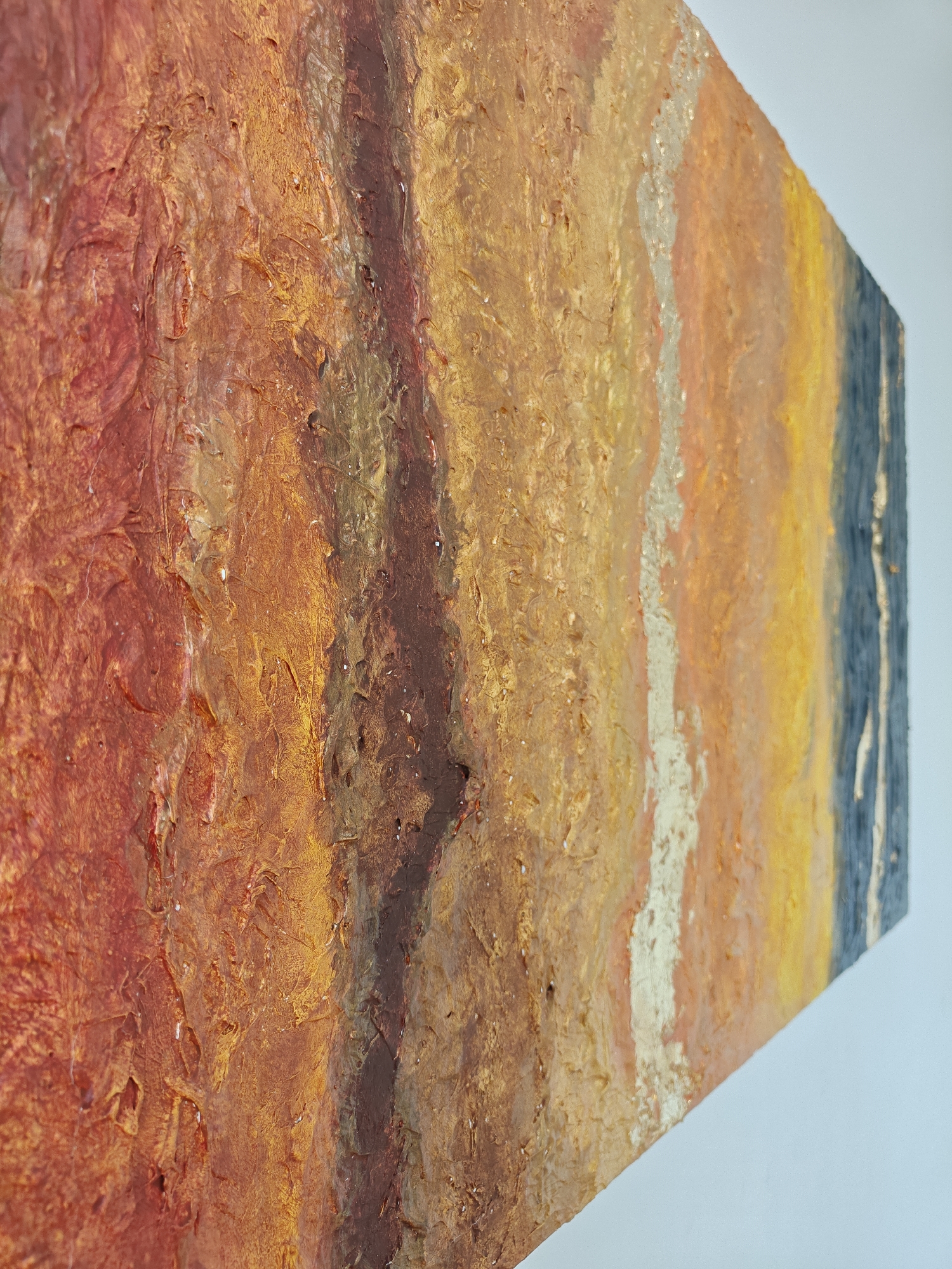 The image shows a close-up of an abstract painting with a heavily textured surface. The painting features a vertical arrangement of colors ranging from deep red on the left, transitioning through orange, yellow, and ending with a dark blue on the right edge. There are hints of gold or silver metallic paint interspersed among the other colors, especially near the center. The painting is mounted on a wall, and the angle of the shot suggests it is viewed from the side. The wall is a neutral white. No people or specific objects related to Mumbai, India are visible.
