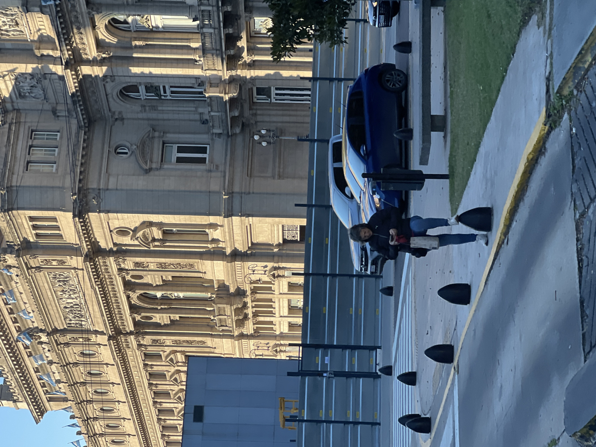 The image depicts a street scene in Buenos Aires, Argentina. The architectural style of the building on the left side of the frame is ornate and classical, possibly European-influenced. The building is beige. Argentine flags are visible on the building's facade. A modern, gray building is in the foreground to the right of the classical building. A woman is walking along the sidewalk. She is holding a phone or a small device in her hand and carrying a white bag. Blue cars are parked along the street. Black bollards delineate the sidewalk. A yellow line is painted along the edge of the curb. The sky is clear and blue. The photograph is taken during daylight hours. The street is lined with tall black posts. A green grassy area is between the cars and the sidewalk. The image is rotated ninety degrees counterclockwise.