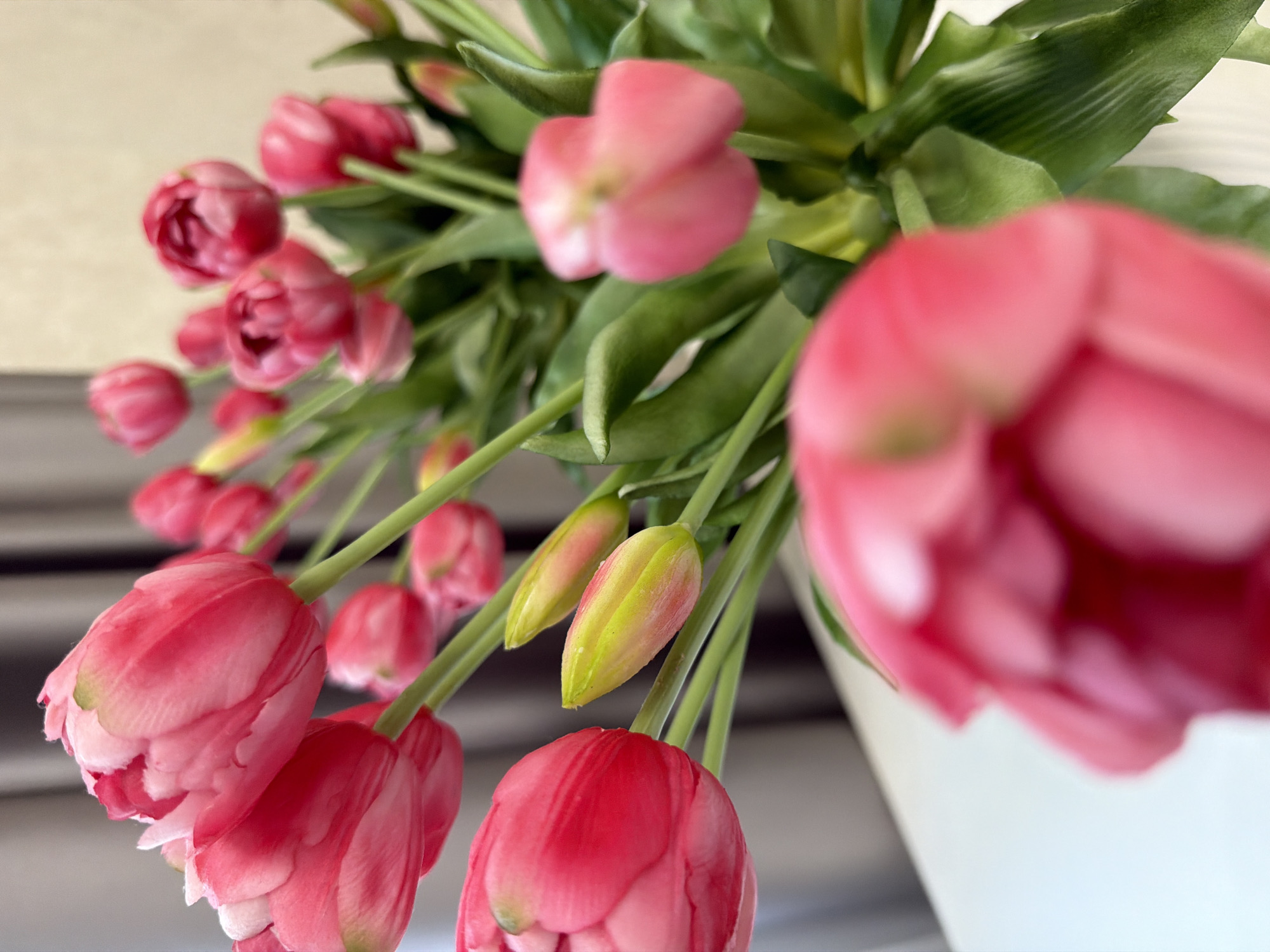 A bouquet of pink tulips is arranged in a vase, resting on a white surface. The tulips are in various stages of bloom, with some fully open and others still in bud. The petals are a vibrant pink, with some lighter pink or white accents. The stems are green and slender, and the leaves are a deep green. The background is blurred, but appears to be a light-colored wall and vertical blinds. The lighting is soft and natural.