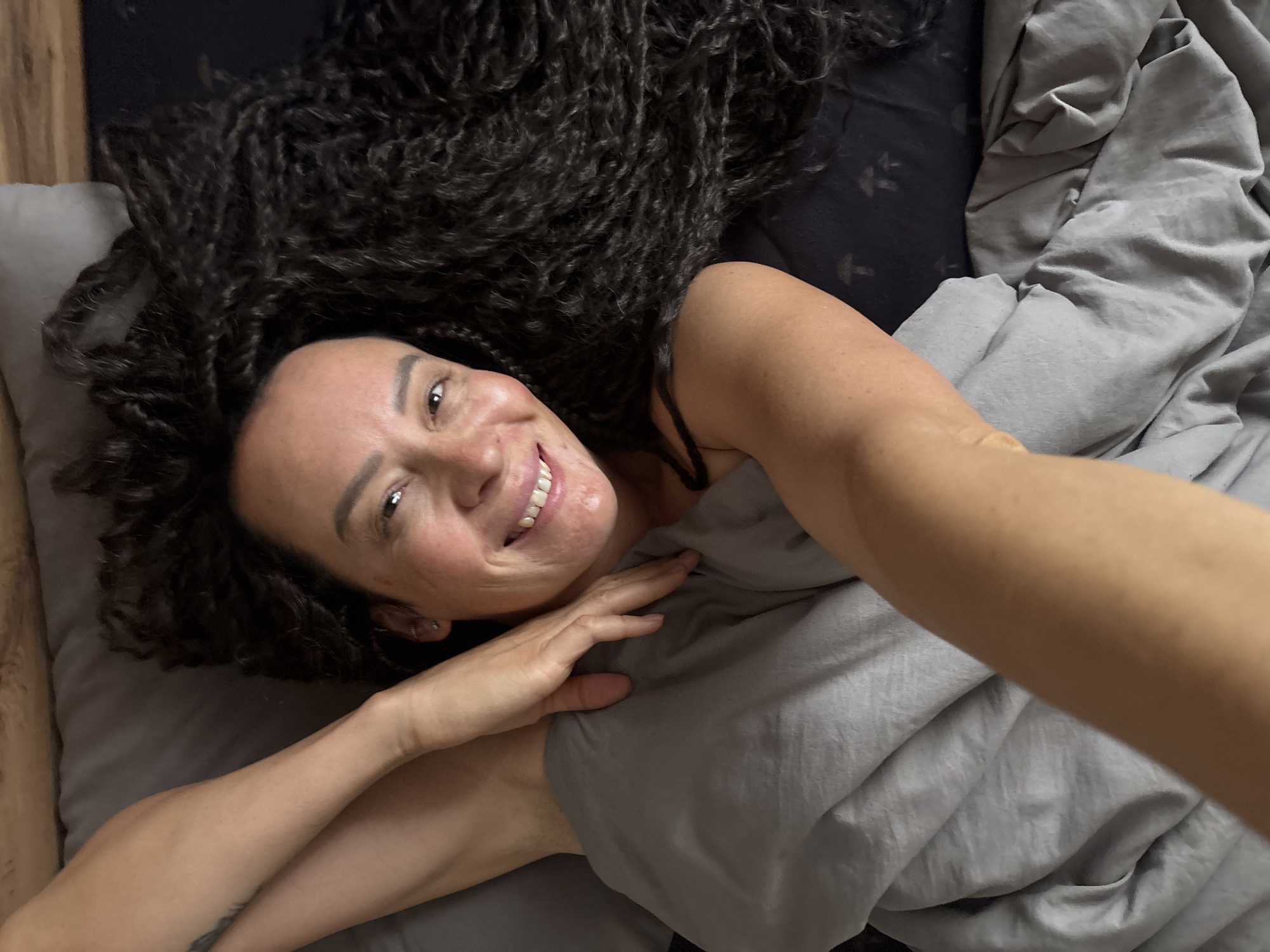 A woman with dark, curly hair is lying on a bed in what appears to be an indoor setting. The bedding includes a dark gray sheet, a light gray duvet, and a light gray pillow. A small portion of a wooden surface is visible in the upper left corner, indicating a headboard or bedside table. The woman is smiling and looking up at the camera, with one arm visible resting on the duvet. The location within Chaikovsky, Russia cannot be determined from the image.