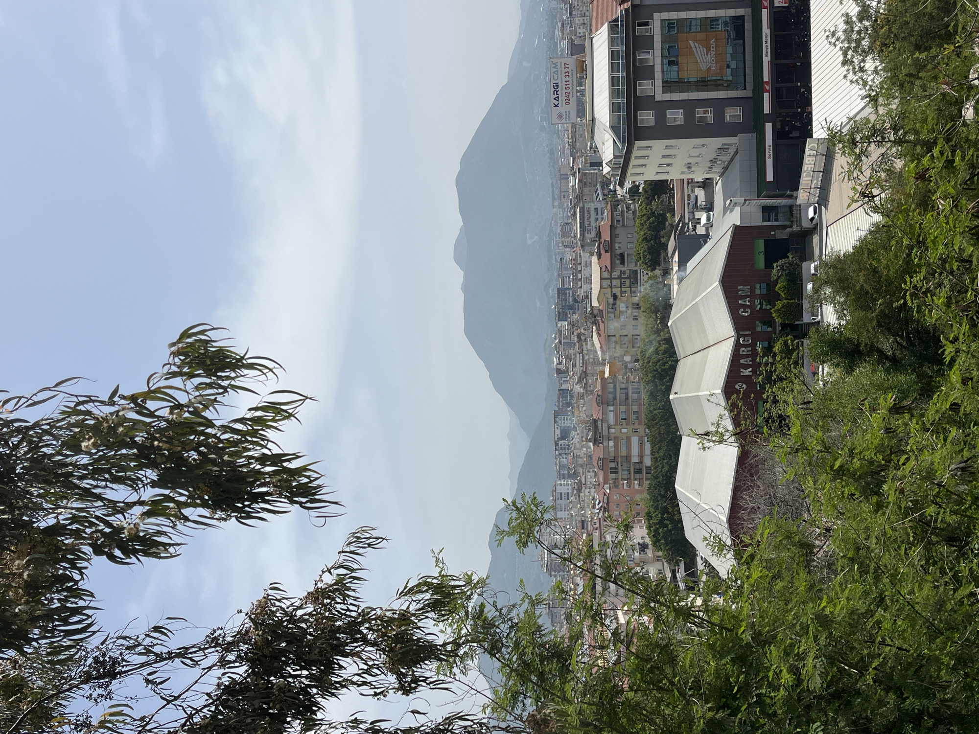 The image shows a slightly elevated view of Alanya, Turkey, with a mountain range visible in the background shrouded in a hazy atmosphere. In the foreground, a cityscape stretches towards the mountains, featuring various buildings, including residential apartments and commercial structures.
The architecture includes modern high-rise buildings, along with older, more compact structures. Vegetation is present in the foreground, particularly trees with dense foliage. A building is labelled "KARGI CAM", and another has a "HONDA" logo.