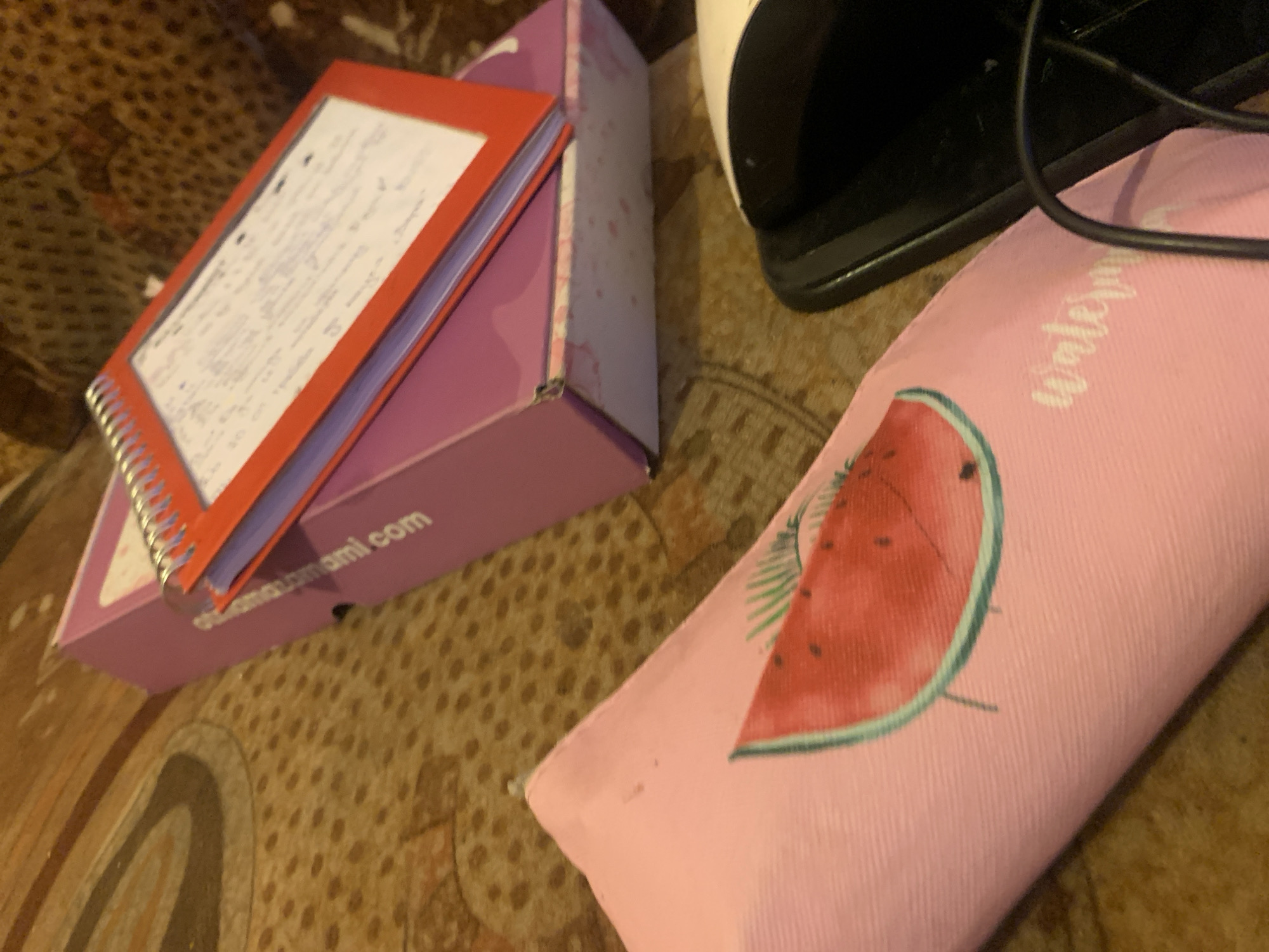The image depicts a flat lay arrangement of items on a patterned surface, possibly a rug or carpet. In the foreground, a pink pencil case adorned with a watermelon slice graphic is partially visible. To the right of the pencil case, a black object, possibly a computer mouse or part of a charging device, is present. In the background, a pink box with a marbled pink and white pattern supports an open red notebook with spiral binding. The notebook contains handwritten notes in blue ink. The overall scene appears to be indoors, in a domestic setting, and there is no indication of its location in Krushev Dol, Bulgaria. The image does not contain any people or any actions taking place.