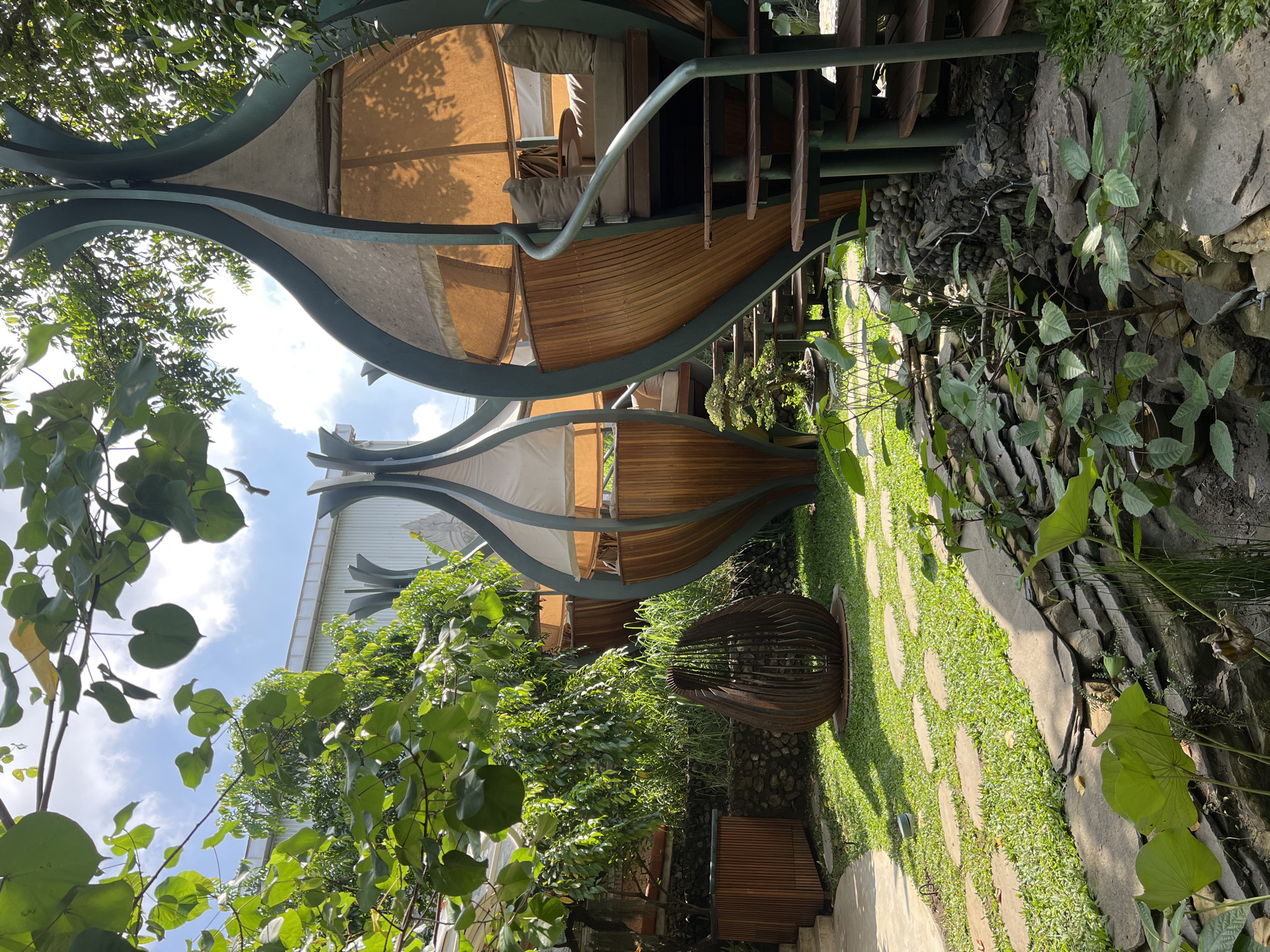 This media file presents a lush, tranquil outdoor scene, likely a resort or garden area in North Kuta, Indonesia. The central subject is an architecturally unique structure featuring organic, wave-like dark green metal supports that frame segments of light-colored fabric awnings and curved wooden panels. This multi-level design includes visible wooden steps and a metal handrail, suggesting an elevated seating or dwelling area nestled within the greenery.
The ground level features a pathway composed of irregular grey stone pavers set within vibrant green grass. This path is bordered on the right by a natural arrangement of rocks and dense foliage, indicating either a dry stream bed or a garden feature. The entire space is enveloped in abundant tropical vegetation, with large green leaves dominating the foreground and various shrubs providing dense cover around the structures.
A large, distinctive sculptural object, resembling an open sphere or ovoid made of stacked dark, horizontal elements, is prominently placed on a grassy patch near the pathway. The scene is illuminated by bright sunlight under a clear blue sky with scattered white clouds, suggesting it is midday or early afternoon, with strong shadows cast across the grass. No people or specific activities are depicted, but the meticulously designed environment evokes a sense of peace and natural harmony. No visible text is discernible in the image.