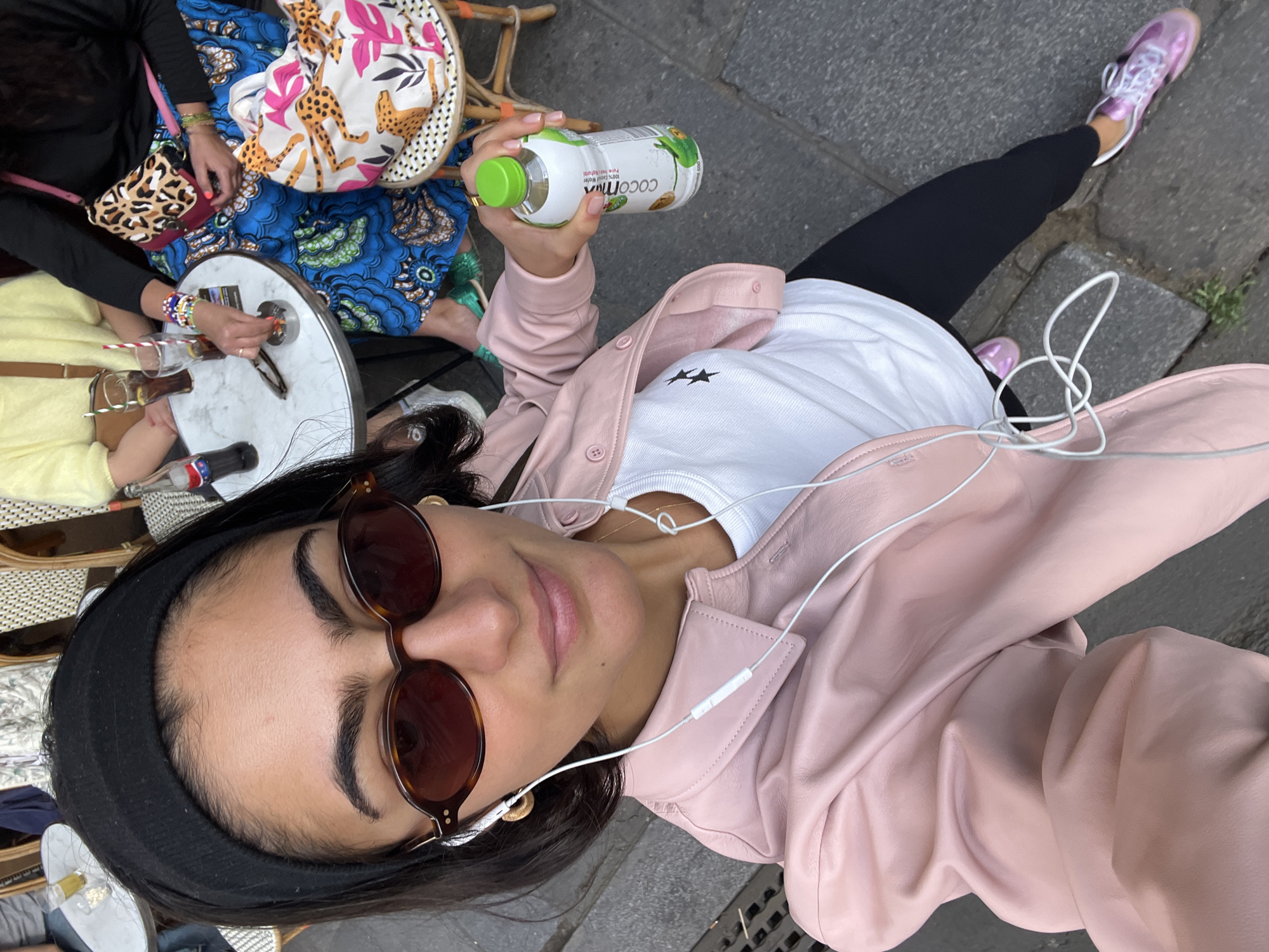 This high-angle selfie captures a casual daytime scene, likely at an outdoor cafe or street-side seating area in Paris, France. The primary subject is a woman taking the photograph, seen from above, wearing a light pink leather jacket over a white t-shirt with a black star logo, black leggings, and pink athletic shoes with white laces. She has dark hair, styled with a black headband, and wears round brown sunglasses. White wired earbuds are visible around her neck and leading to her ears.
She holds a bottle of "Cocomilk" coconut water in her right hand, with "100% Cocomilk Water" and "Pure natural water" printed on the label, alongside a green frog illustration.
To her left, other individuals are partially visible around a small, round white marble-effect table. One person is wearing a yellow top and colorful beaded bracelets, their hand reaching towards a drink. Another person is seen with a blue patterned garment and a leopard print bag. On the table are two small bottles, possibly Coca-Cola, each with a striped straw (red and white, blue and white).
The ground is paved with grey cobblestones or similar urban-style pavers. The overall atmosphere is relaxed and social, depicting friends enjoying a break outdoors.