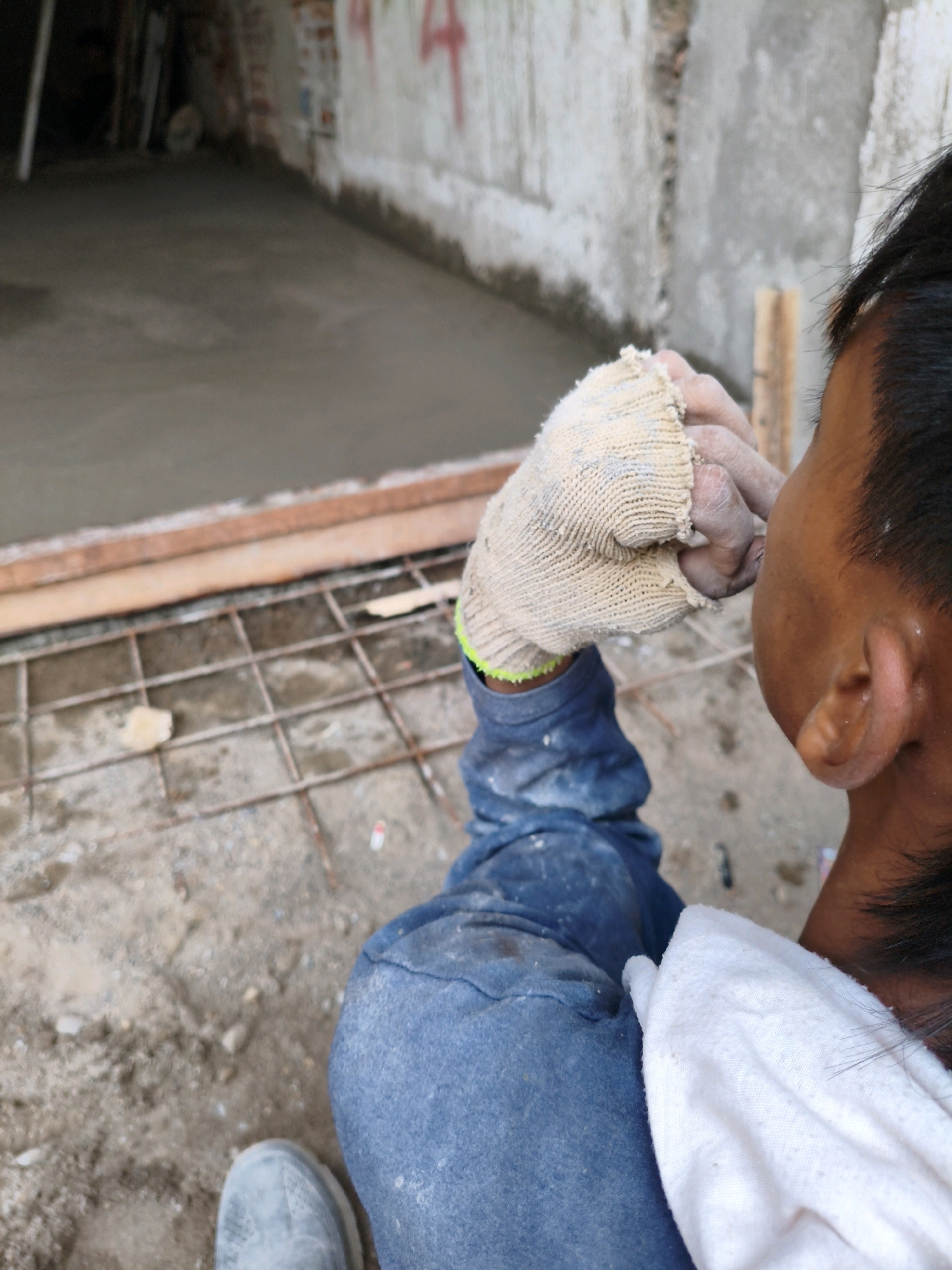 The image shows a close-up view of a construction worker's gloved hand near their face. The worker is wearing blue denim pants and a white cloth is draped over their shoulder. The background suggests an outdoor construction site with a concrete floor, rebar, and unfinished walls. Red graffiti is visible on the wall in the background. The scene appears to be in Bekasi, Indonesia, given the context.