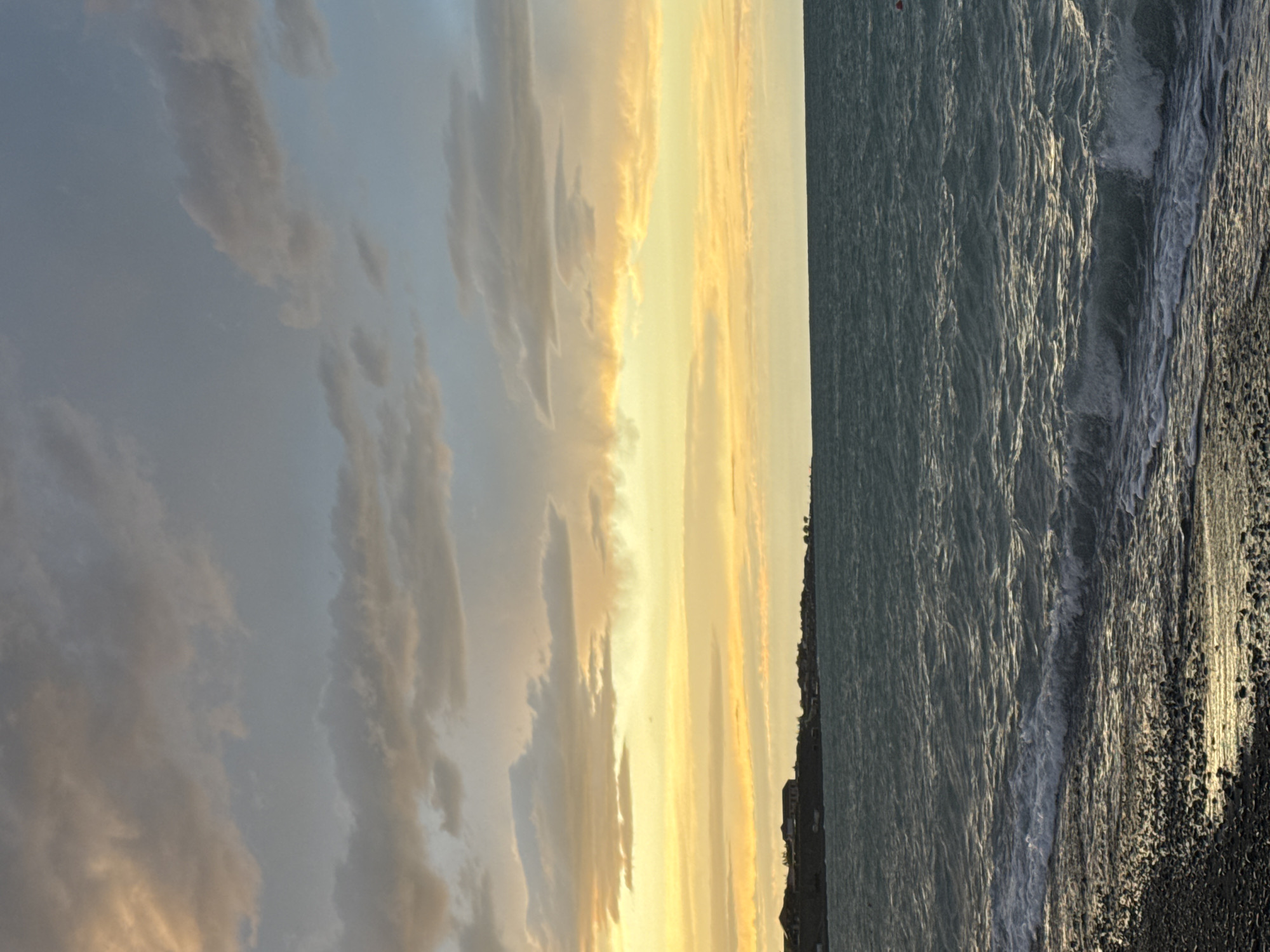 The image depicts a serene coastal scene at dusk in the city of Akshi, Kazakhstan. The sky is filled with dramatic clouds, displaying a gradient of colors from a pale yellow near the horizon to a muted gray and blue higher up. The sun is setting, casting a warm glow across the water and the sky.
In the foreground, a rocky beach is visible, with small waves gently lapping at the shore. The water is a deep blue-green, with white foam from the breaking waves creating a dynamic contrast against the darker water.
In the distance, along the coastline, a line of buildings can be seen. These structures appear to be houses or low-rise commercial buildings, nestled along the edge of the land. The overall impression is one of tranquility and natural beauty, characteristic of a coastal town.