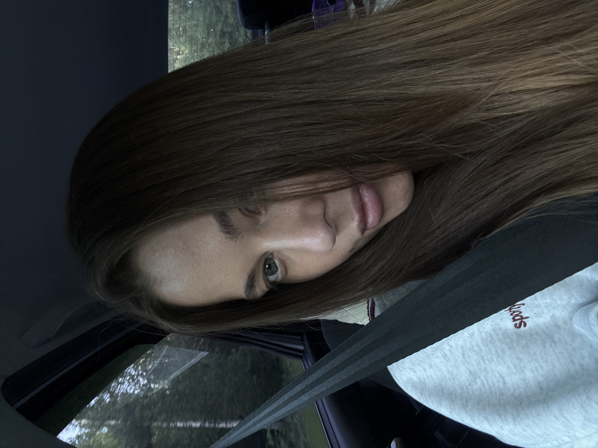 A young woman with long brown hair is in the passenger seat of a car, looking towards the camera. She is wearing a light gray sweatshirt with the word "sporty" embroidered in red on the left chest. A black seatbelt is diagonally across her chest. The window to her left shows a blurred green, wooded environment, suggesting the car is in motion. The interior of the car, including the dark headliner and seat, is visible. The lighting is dim, likely from the late afternoon or early evening. The exact location within Russia is not determinable from the image.