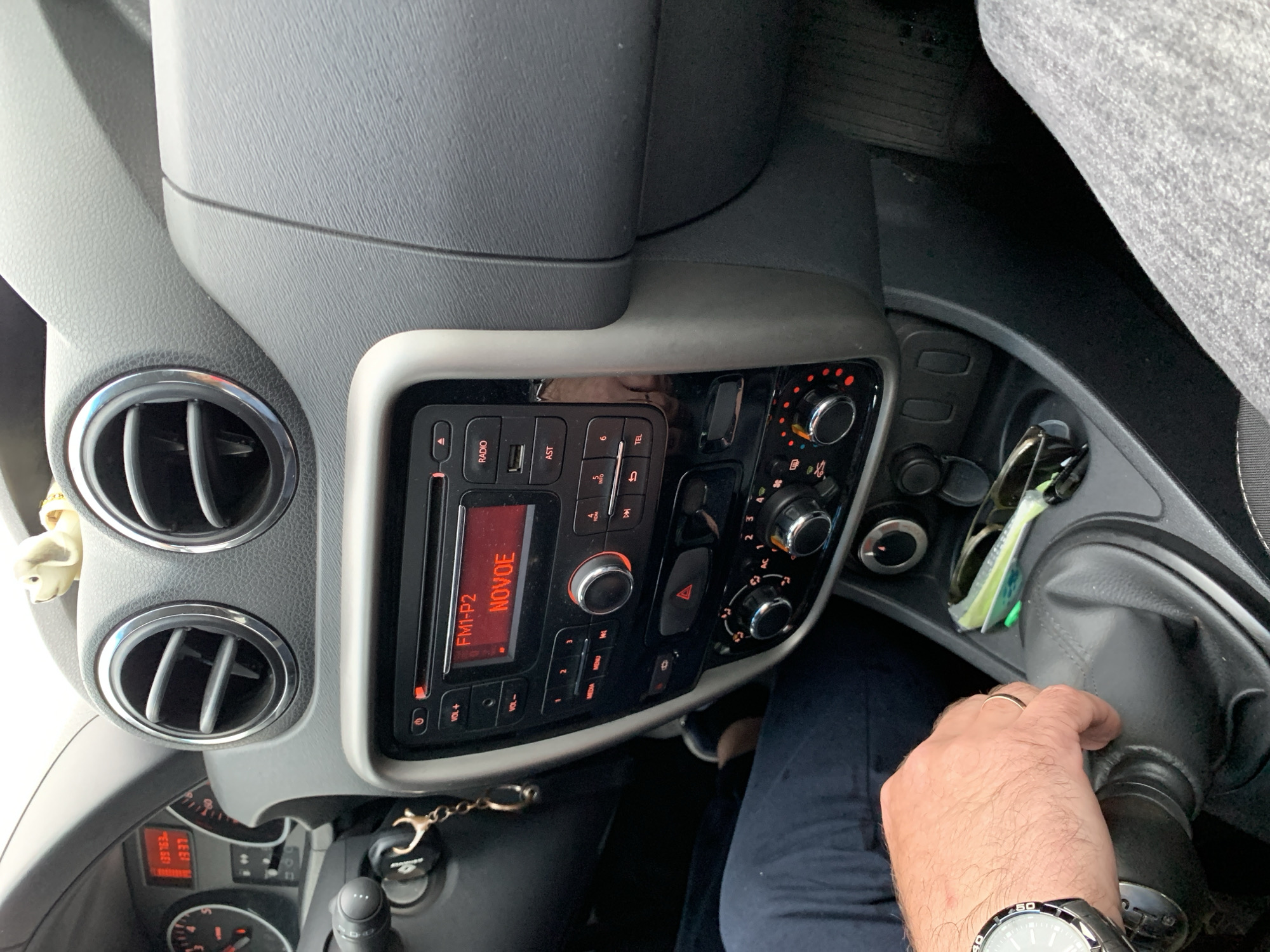 The image shows the interior of a car. A person's hand, wearing a watch and a ring, is on the gear shift. Sunglasses and a key fob are visible in the center console. The car's radio is on, displaying "FM1-P2 NOVOE". The dashboard includes air vents and instrument clusters. The environment appears to be a city in Belarus, possibly Курапольскі сельскі Савет. The image is taken from the perspective of a passenger or the driver.