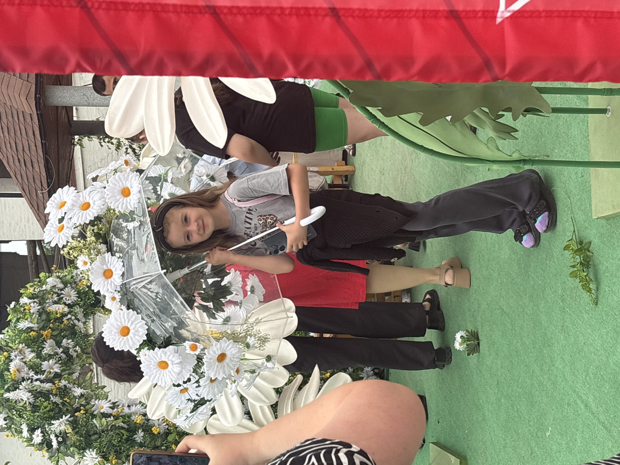 A young girl is standing on green Astroturf, holding a clear umbrella decorated with large white daisies. She is wearing a grey t-shirt with "CREATIVE" printed on it, dark pants, and colorful Crocs. Behind her, a woman in a red dress and another person in black pants are visible, as well as a large floral archway and decorative white daisies. To the right, a bright red banner partially obscures the scene. The location is outdoors, possibly a park or event space, and it is daytime.