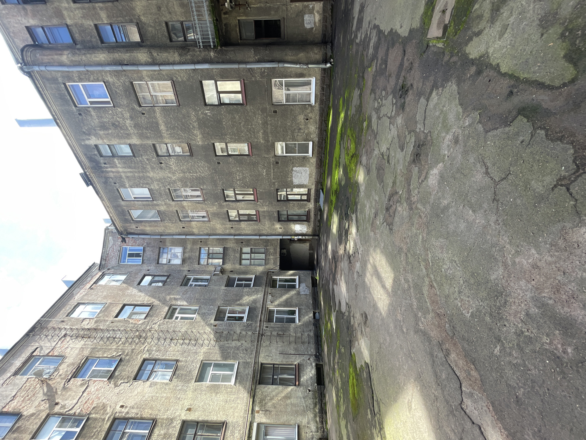 The image captures a view of a dilapidated courtyard in Vyborg, Russia. The courtyard is paved with cracked asphalt and shows patches of moss and grass growing through the pavement. The surrounding buildings are multi-story, with weathered facades and numerous windows. Some of the windows are modern, while others appear older. There is a fire escape on one of the buildings. The sky is visible above the buildings, suggesting it is daytime. The scene conveys a sense of neglect and urban decay.