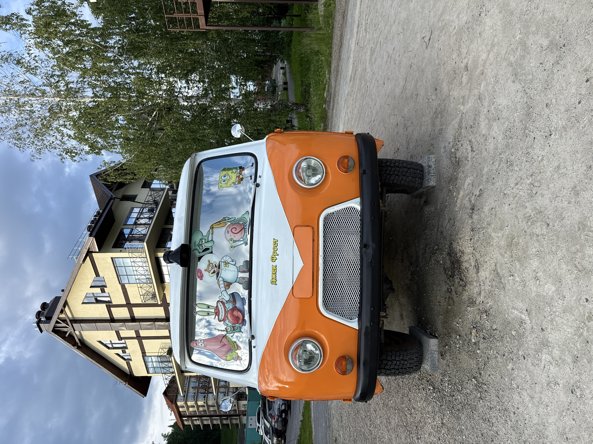 An orange and white van is parked on a gravel surface in Syrostan, Russia. The van is decorated with several SpongeBob SquarePants characters on its windshield. Behind the van, a multi-story building with balconies and dark brown trim is visible. Trees with green foliage stand to the right of the van and building, and a cloudy sky is overhead. The van's front wheels are elevated on rectangular concrete blocks.